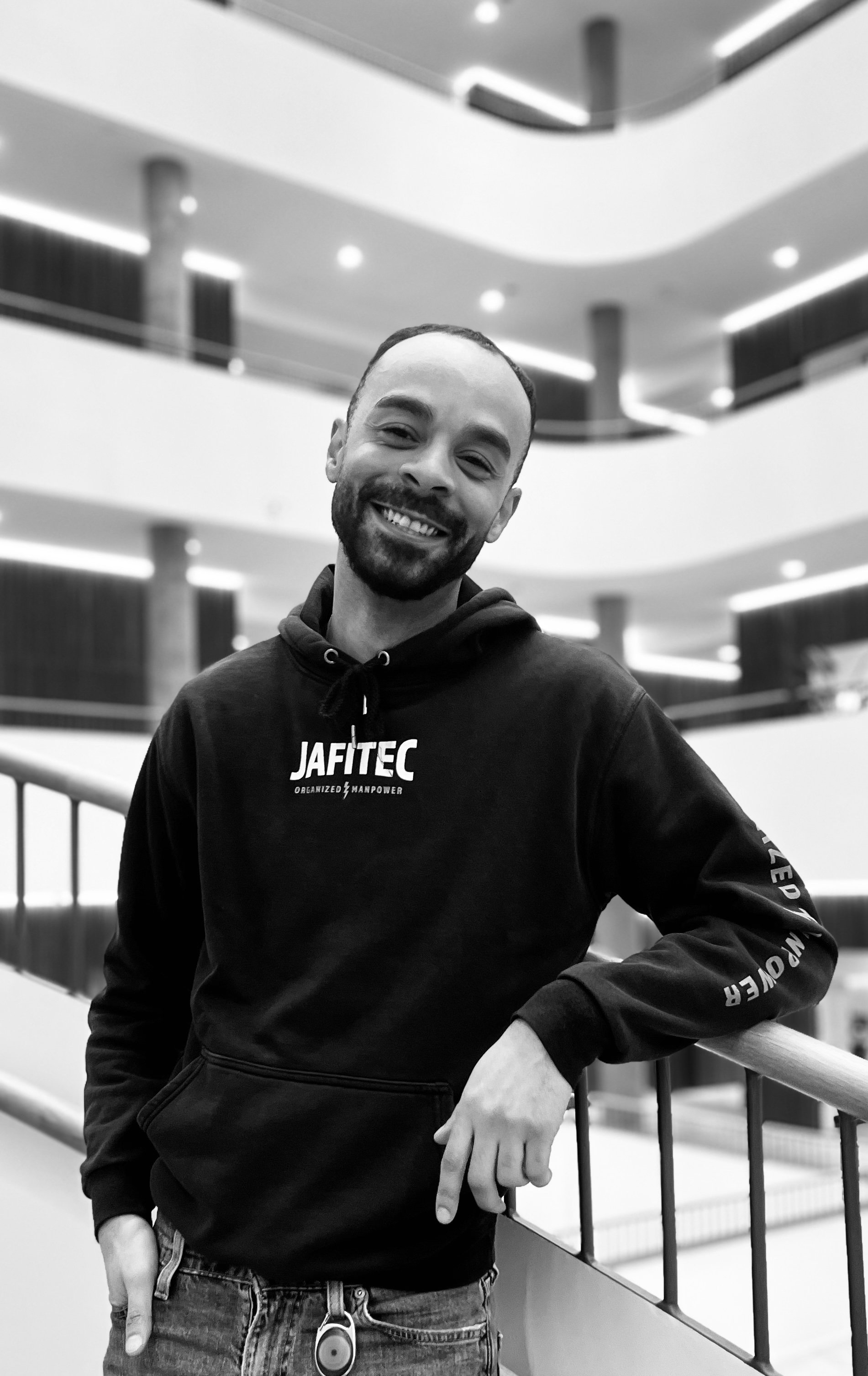 Man wearing a safety helmet, a jacket with reflective stripes, holding a clipboard, standing against a plain wall.