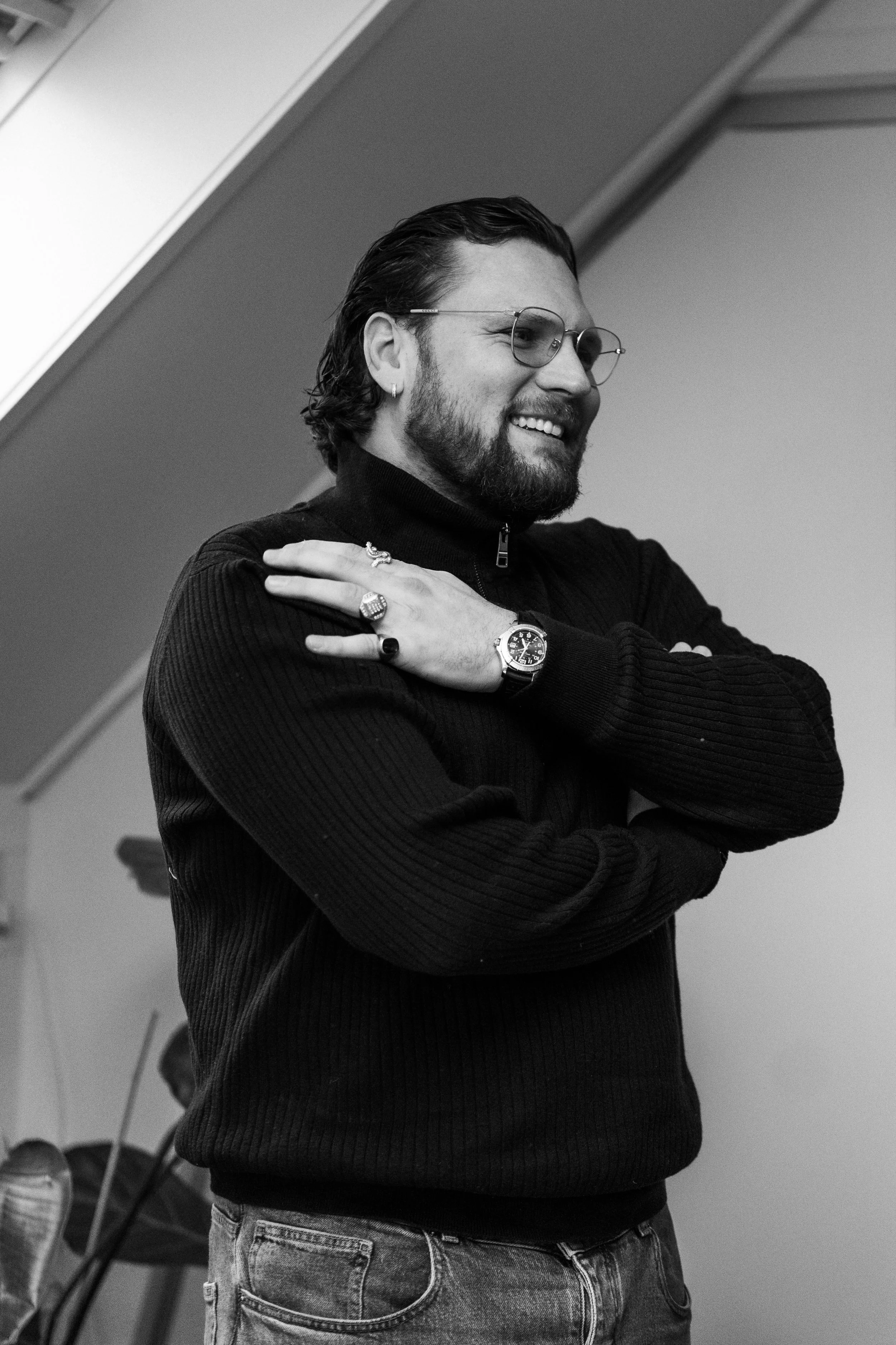 Man standing on a stairway inside a modern building, wearing a reflective safety vest, holding a helmet and transparent glasses, with long hair and a beard, in black and white.