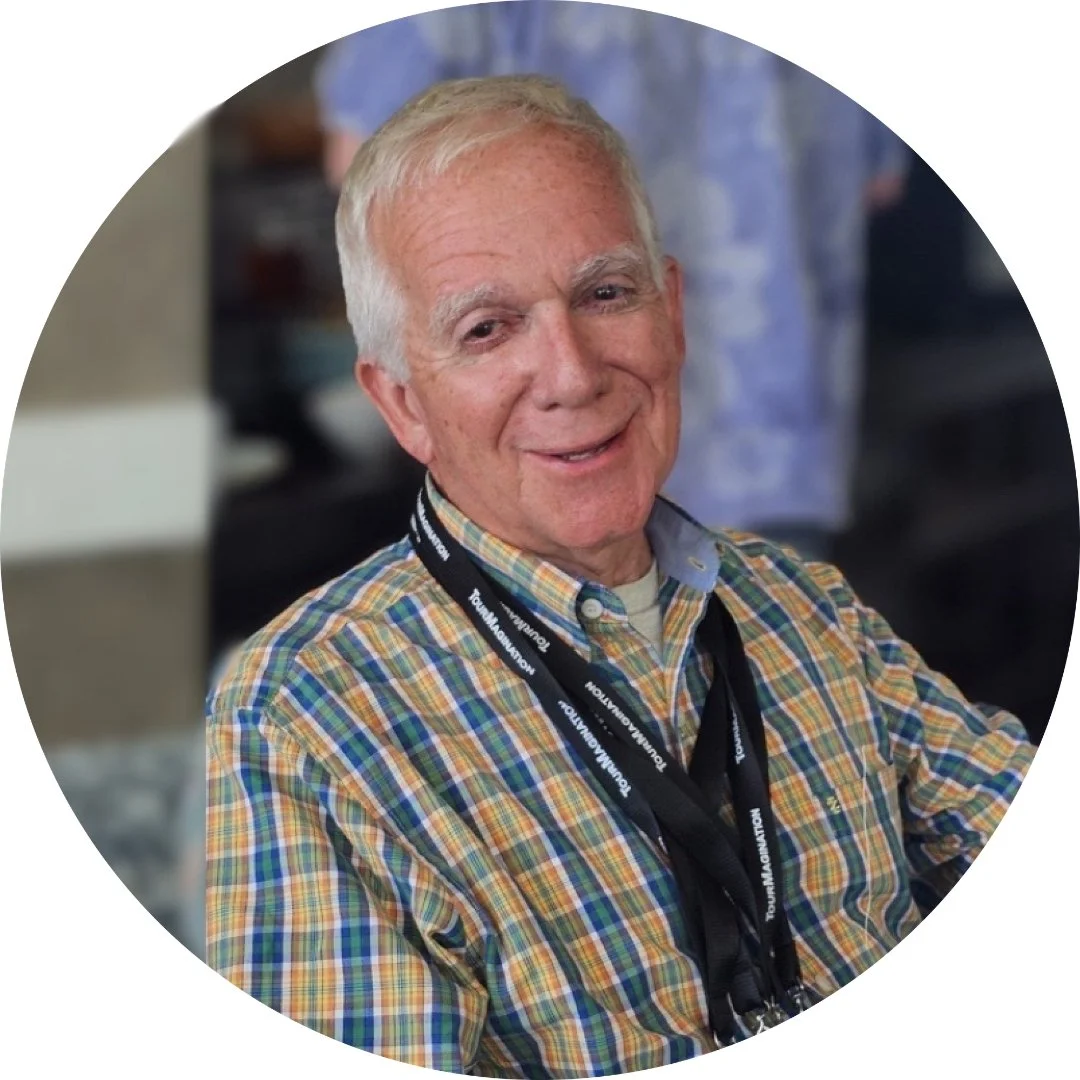 Older man with short white hair smiling, wearing a blue and white checkered shirt, outdoors with green foliage in the background.