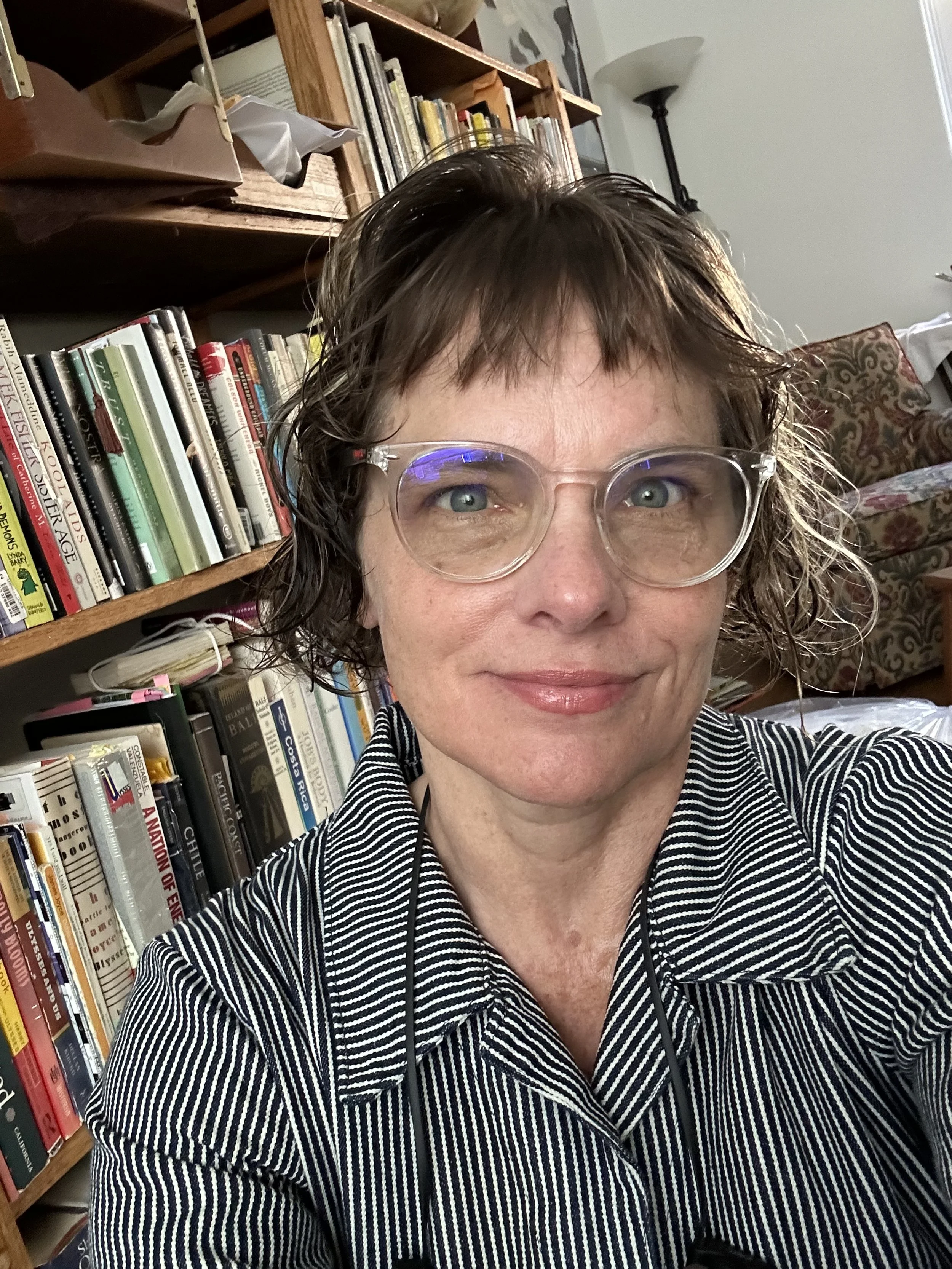 A woman with short, wavy brown hair and glasses taking a selfie in a room with a bookshelf and lamps in the background.