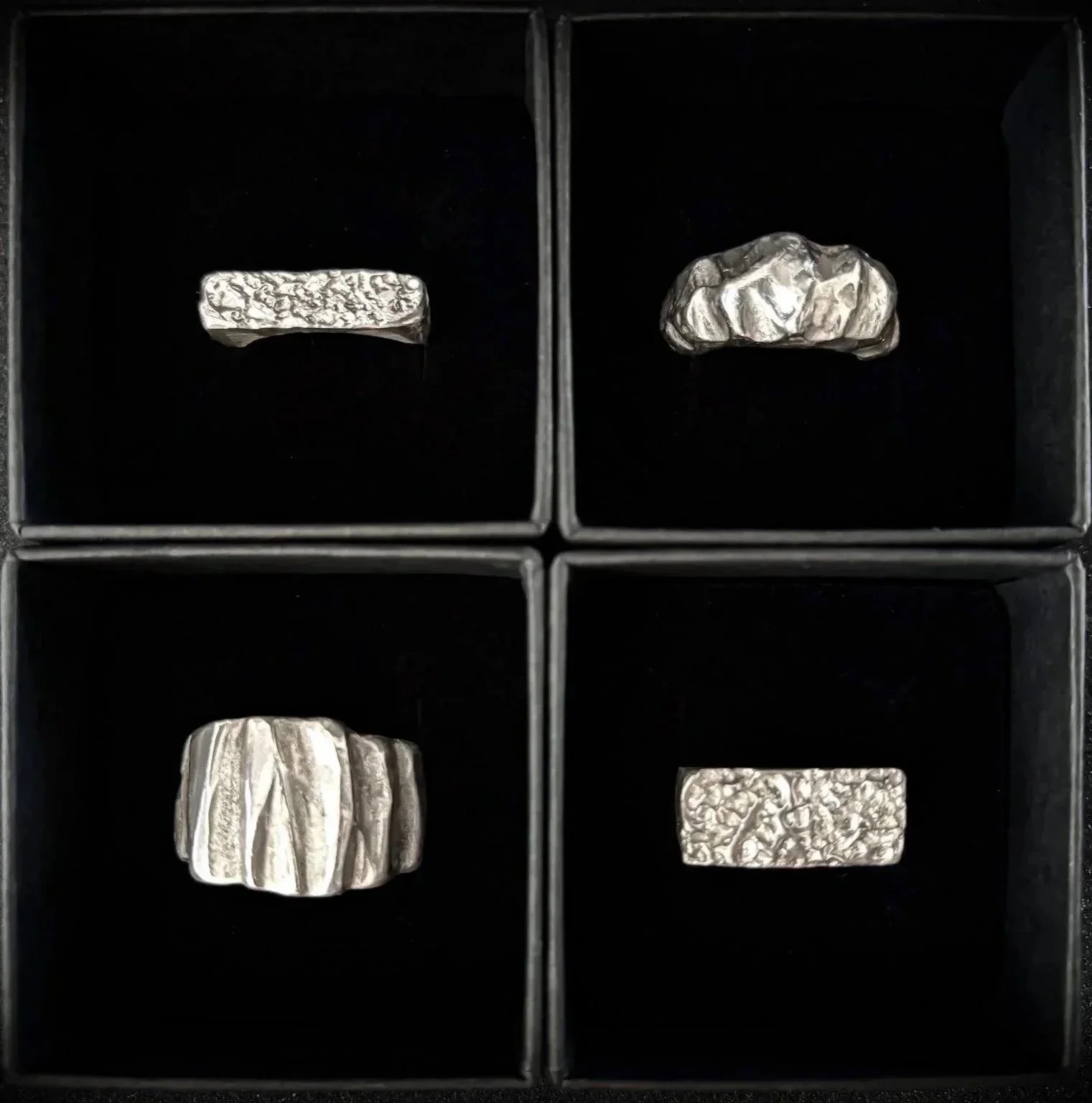 Four hand sculpted silver rings with textured surfaces, each placed in its own black box compartment. The rings are cast in solid 925 and vary in shape and design.