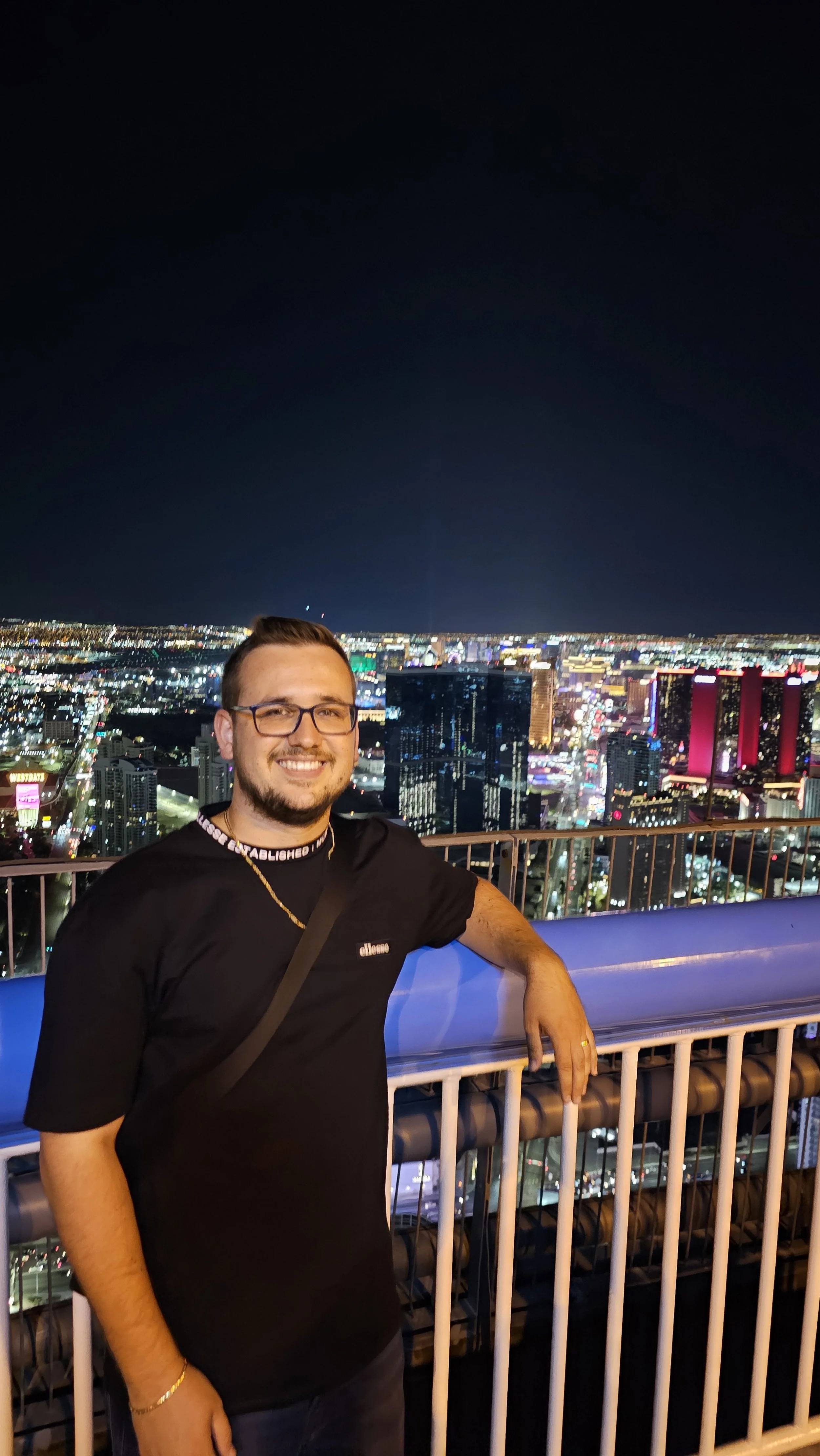 Ein Mann lächelnd auf einer Aussichtsplattform bei Nacht mit einer leuchtenden Stadt im Hintergrund.