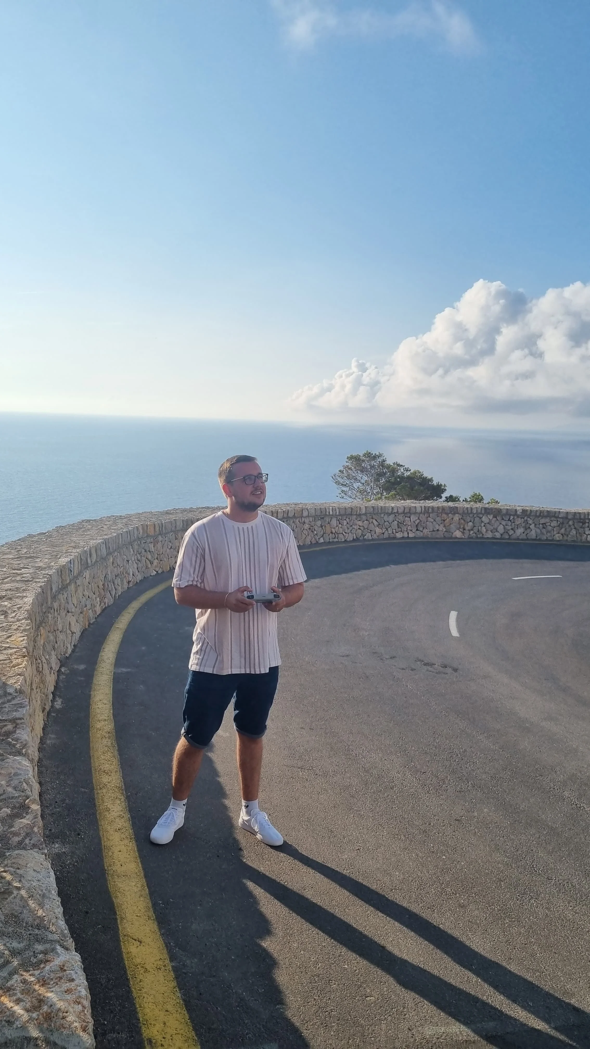 Junger Mann mit Brille, gestreiftem T-Shirt, Shorts und weißen Schuhen steht auf einer kurvigen Straße am Meer mit Blick auf den Himmel und Wolken im Hintergrund.