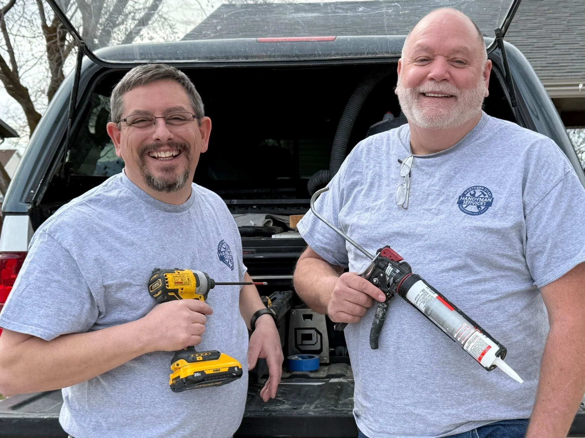 Two men, Jim and Bruce, in gray T-shirts smiling and holding tools, standing behind a work truck, likely working on a project together.