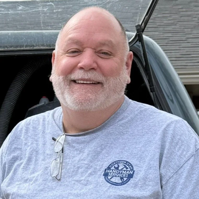 A smiling man with a beard, Bruce, with glasses hanging from the shirt collar of his gray t-shirt with a logo that reads 'Northern Utah Handyman Services', standing outdoors with a house and a vehicle in the background.