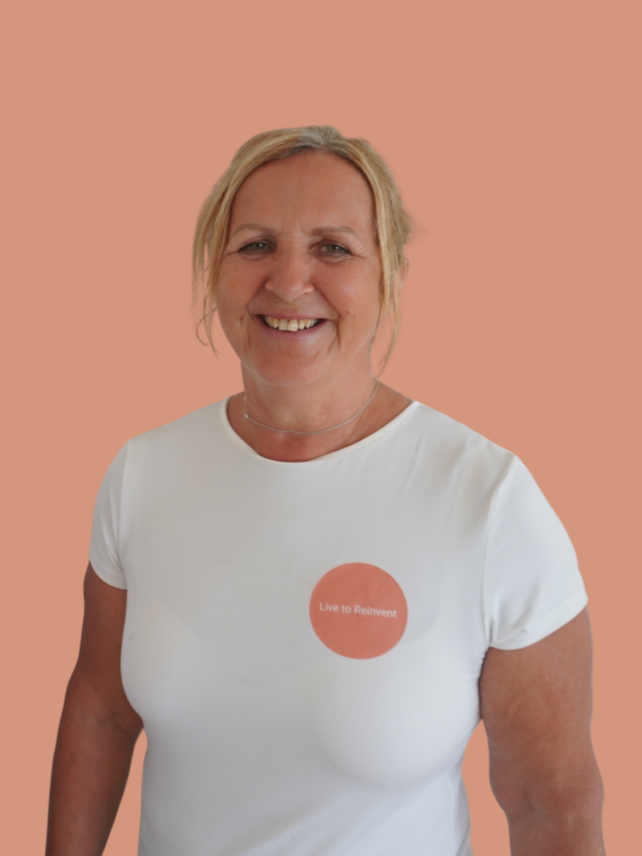 A young woman smiling in front of a plain peach-colored background, wearing a white t-shirt and a necklace.