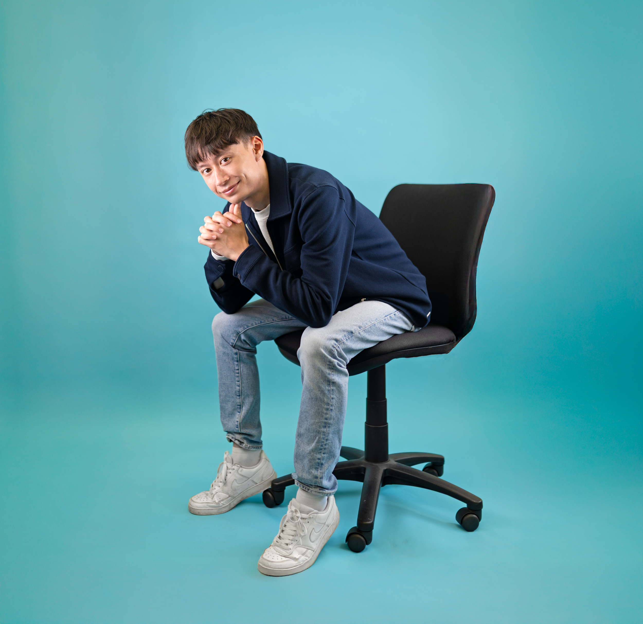 Young man in casual clothing sitting on a black office chair against a teal background, leaning forward with hands clasped, looking at the camera with a slight smile.