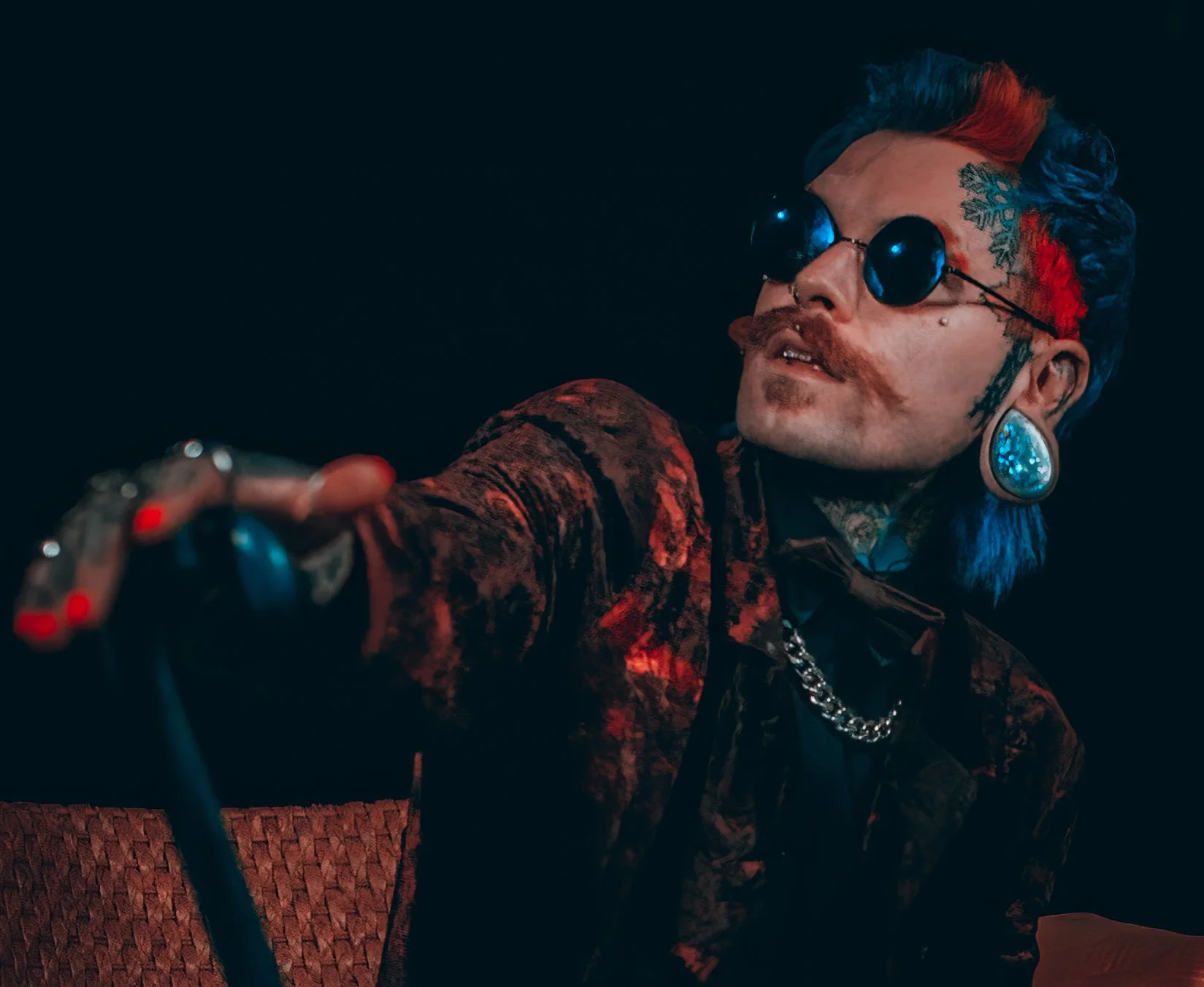A person with multi-colored hair, wearing large round sunglasses and elaborate jewelry, looking to the side in a dark setting.