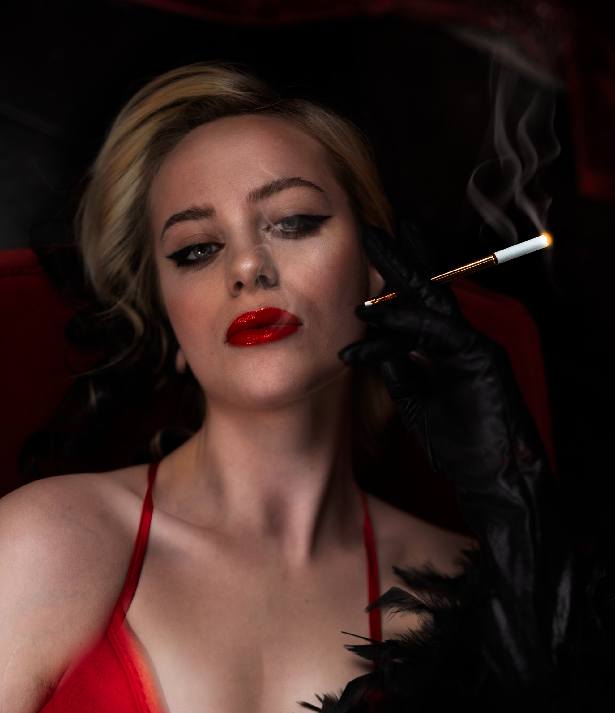 A woman with blonde hair and red lipstick lying down, holding a lit cigarette holder with smoke, wearing a red dress and black gloves, with dark lipstick and eye makeup.