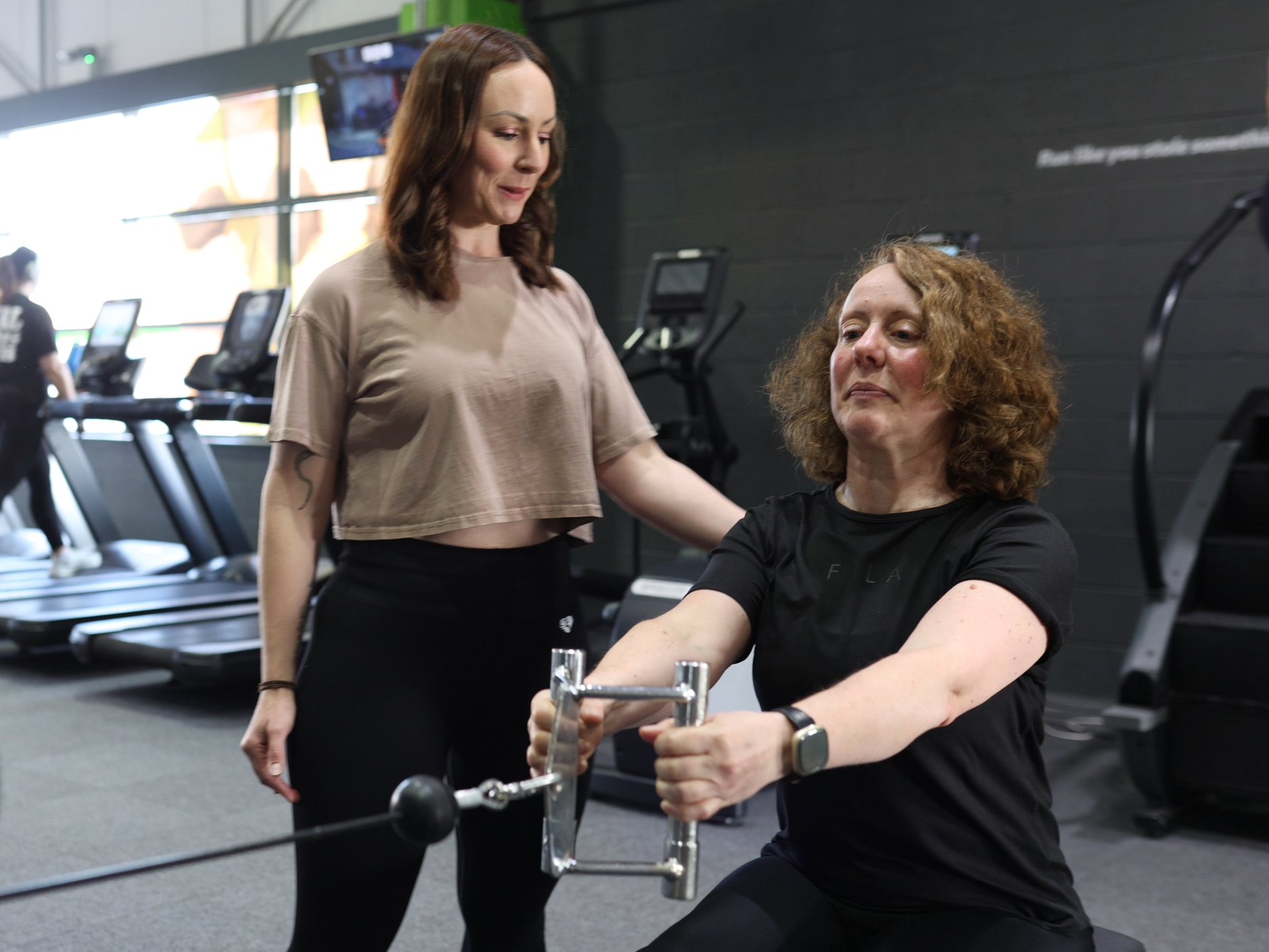 Unposed fitness photography of personal trainer assisting a female client using a cable machine in a gym.