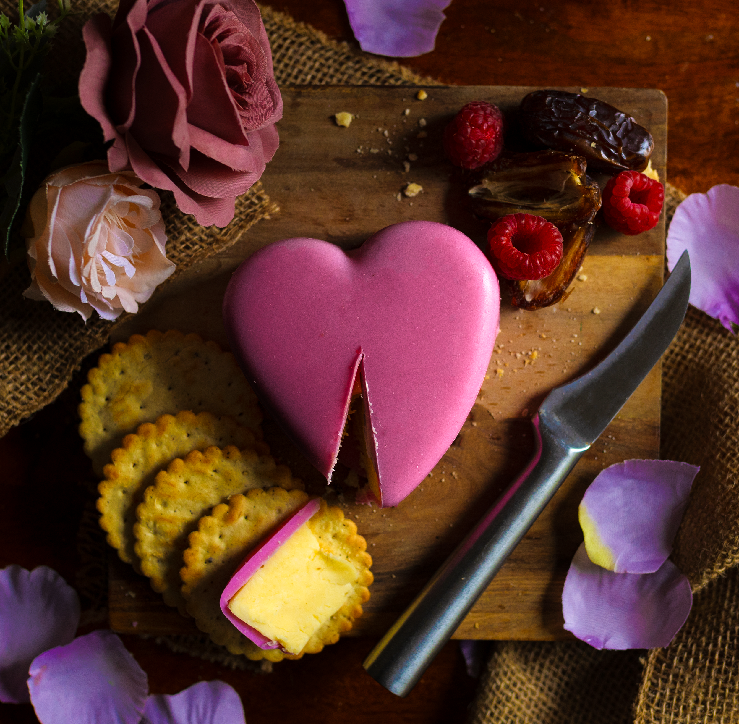 A pink heart-shaped cake with a slice cut out, surrounded by cookies, raspberries, and dates, on a wooden cutting board with flowers and petals around.