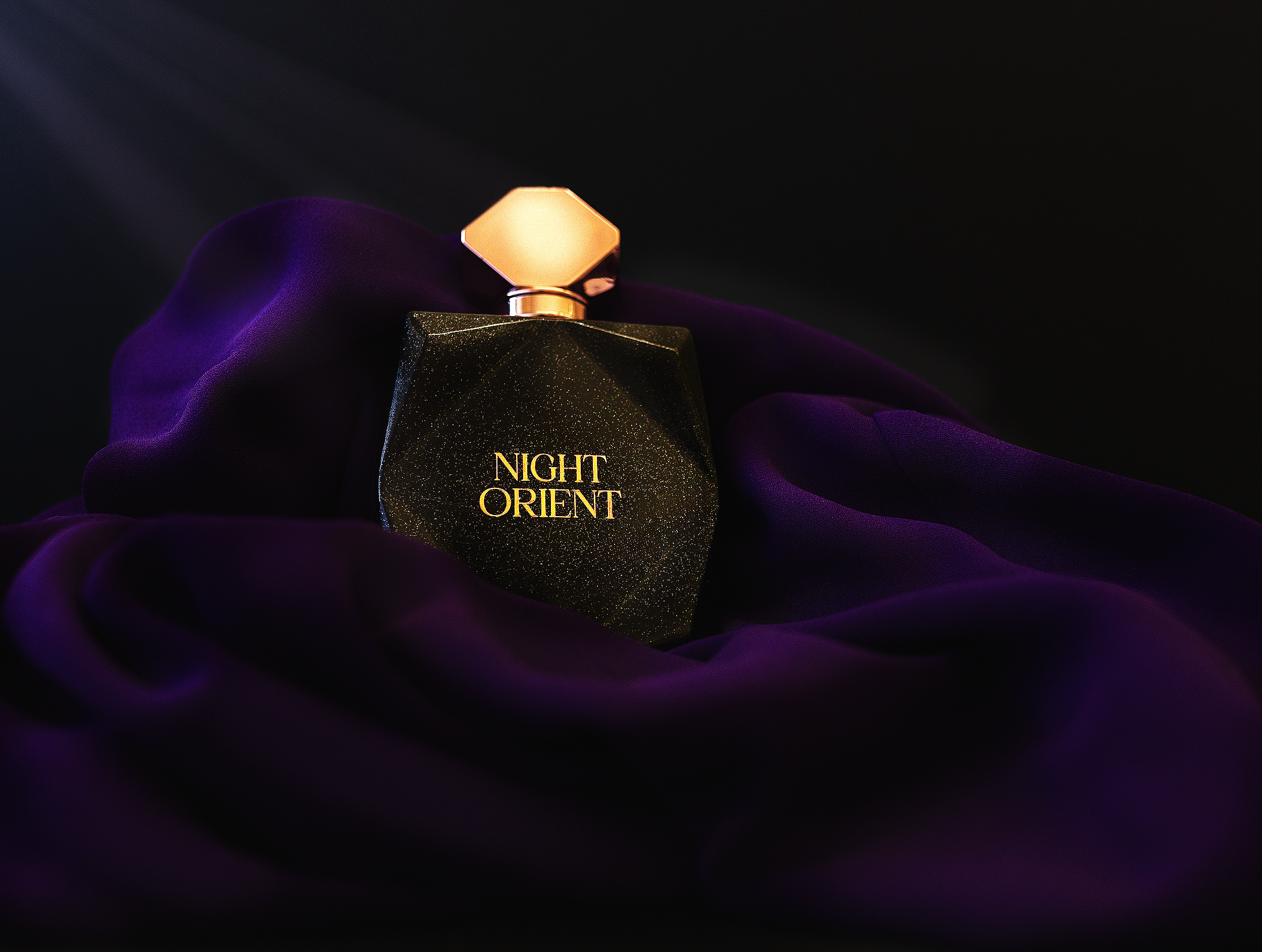 A black perfume bottle with gold specks, labeled 'Night Orient,' resting on deep purple fabric with a gold cap and in a dark setting.