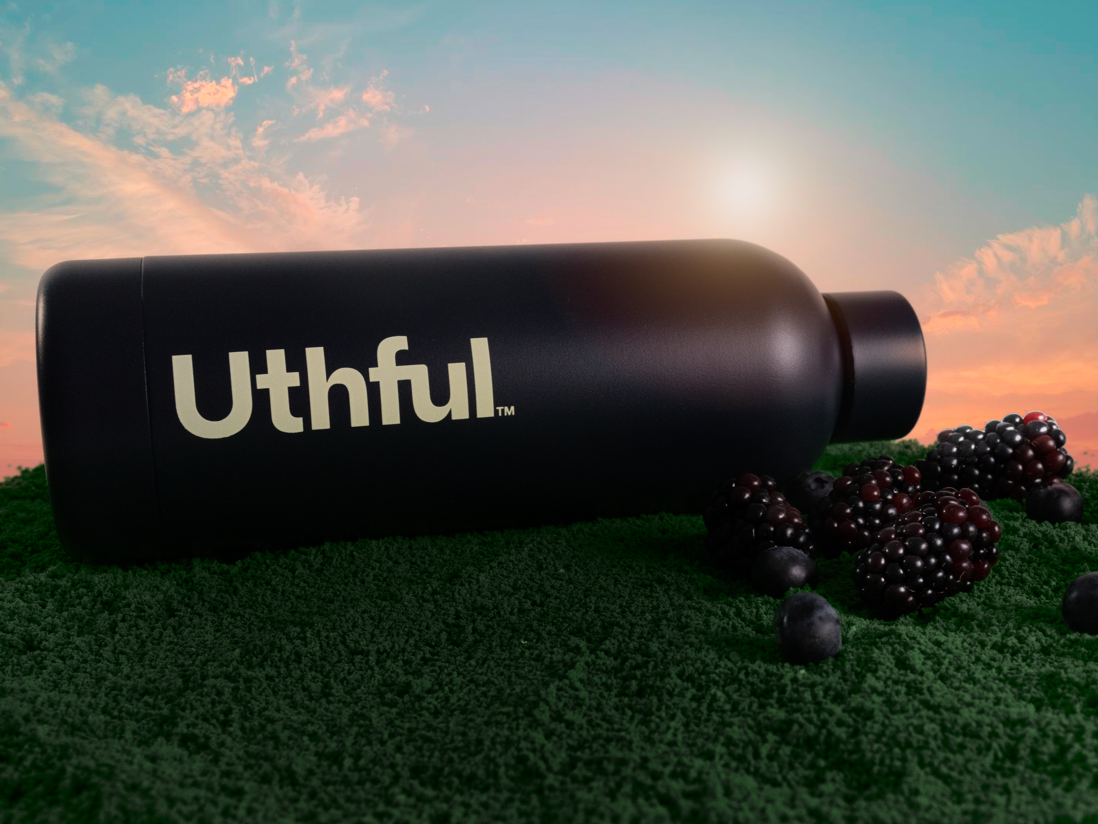 Surreal oversized still life image a black bottle with the word 'Uthful' on it, lying on green grass with blackberries around it, with a sunset sky in the background.
