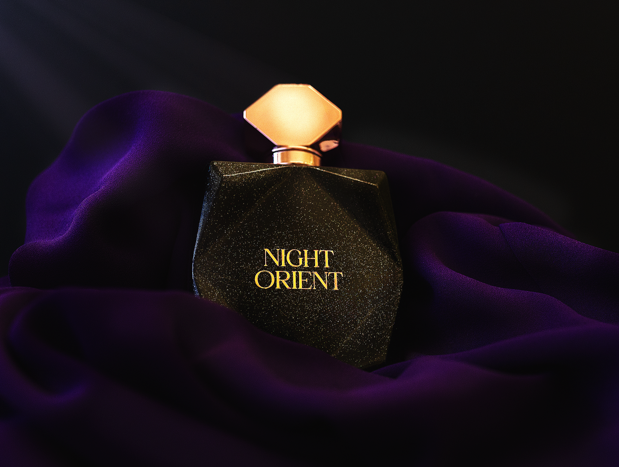 A black perfume bottle with gold speckles and gold lettering that reads 'Night Orient,' resting on purple velvet fabric with a dark background.
