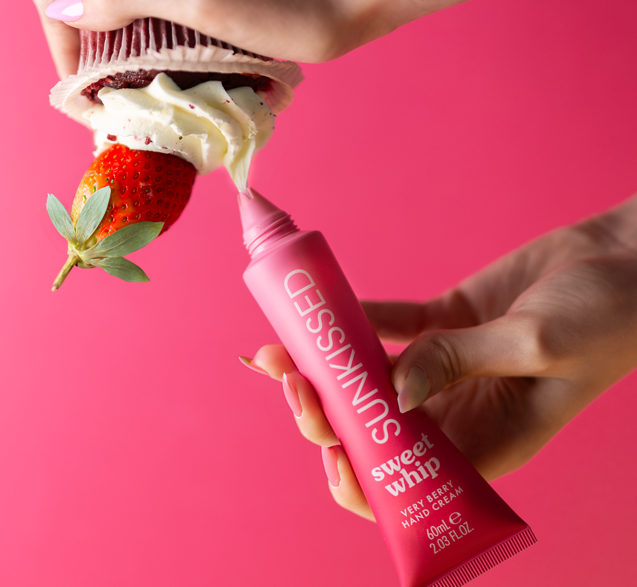 A quirky commercial cosmetics photo person holding a pink squeeze tube labeled 'Sweet Whip' strawberry hand cream, applying it to a strawberry and whipped cream dessert against a pink background.