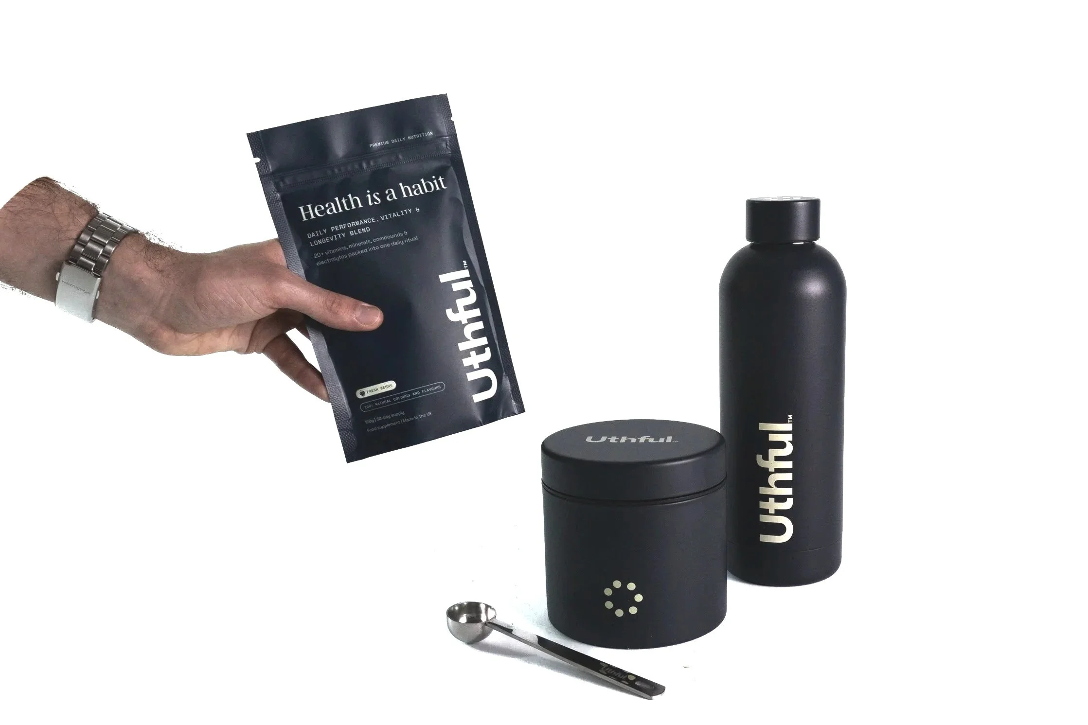 A ecommerce product photo showing a person holding a black packet labeled 'Health is a habit' with three black wellness products branded 'Uthful': a small jar, a tall bottle, and a spoon, all against a white background.