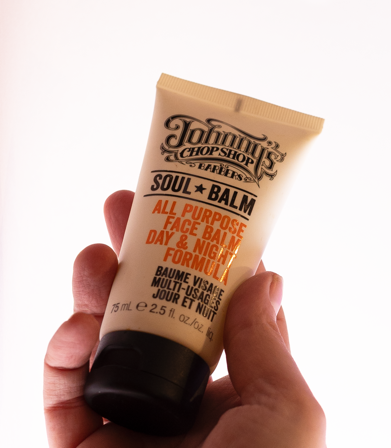 A hand holding a tube of Johnny's Chop Shop Soul Balm, designed for face application, with a light background.