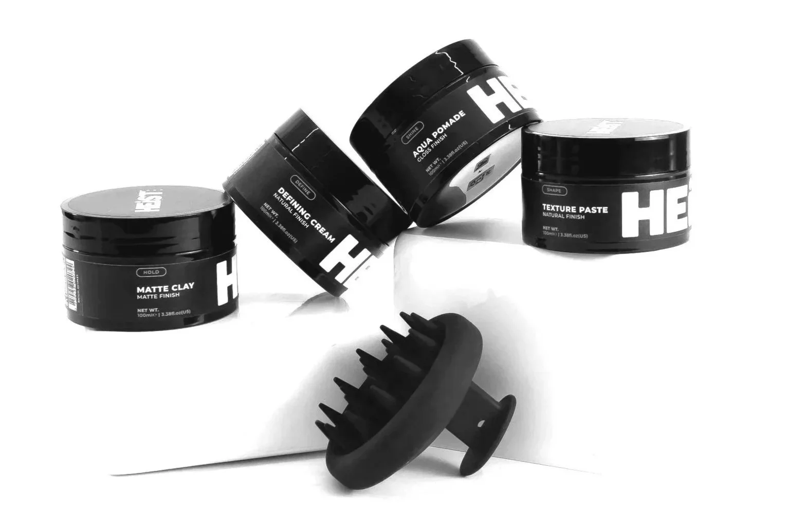 Set of black hair styling products including matte clay, defining cream, aqua pomade, and texture paste, with a black scalp massager in front, arranged on a white surface.