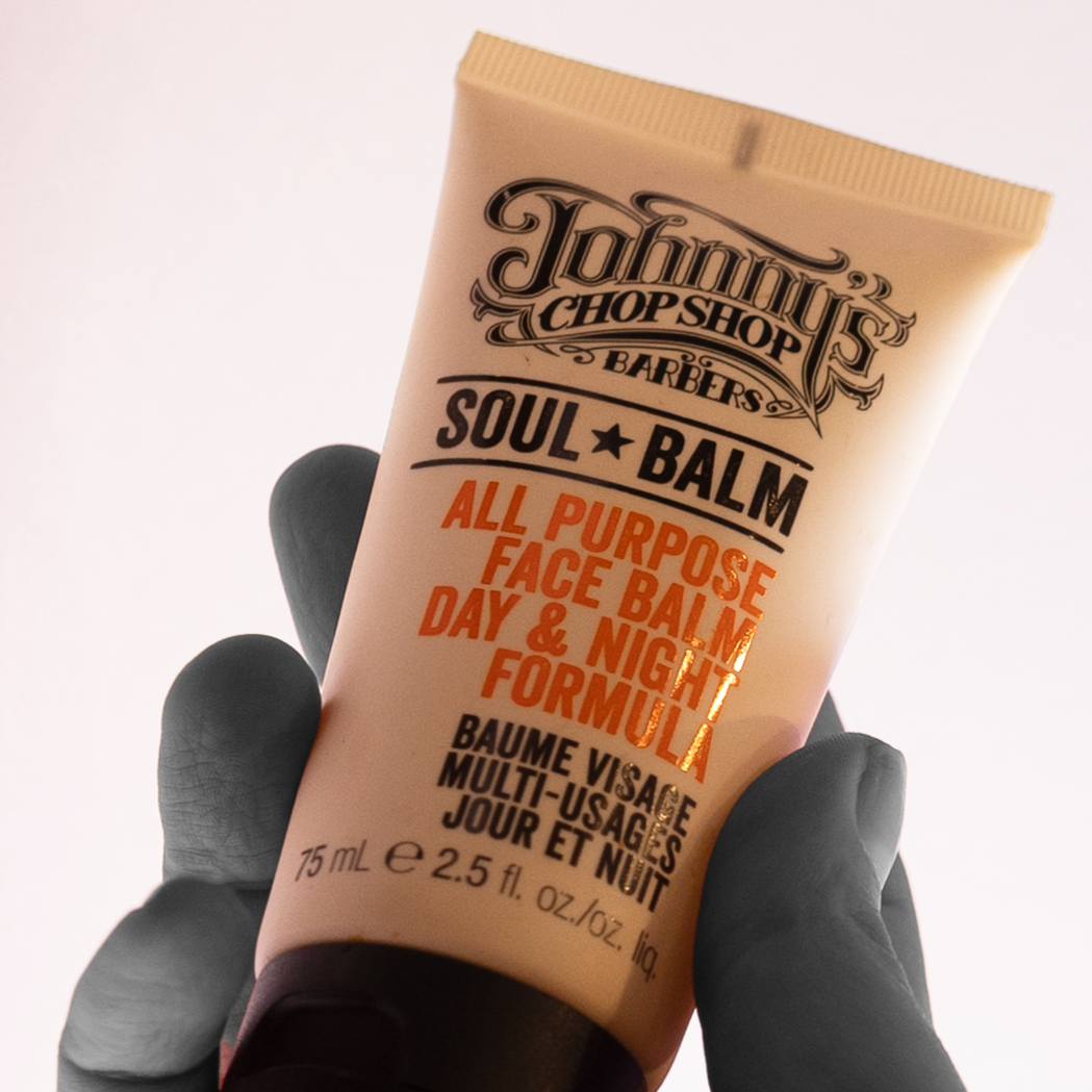 A hand holding a tube of Johnny's Chop Shop Soul Balm, a multi-purpose face balm for day and night use, with black gloves on. The tube is beige with black and orange text.