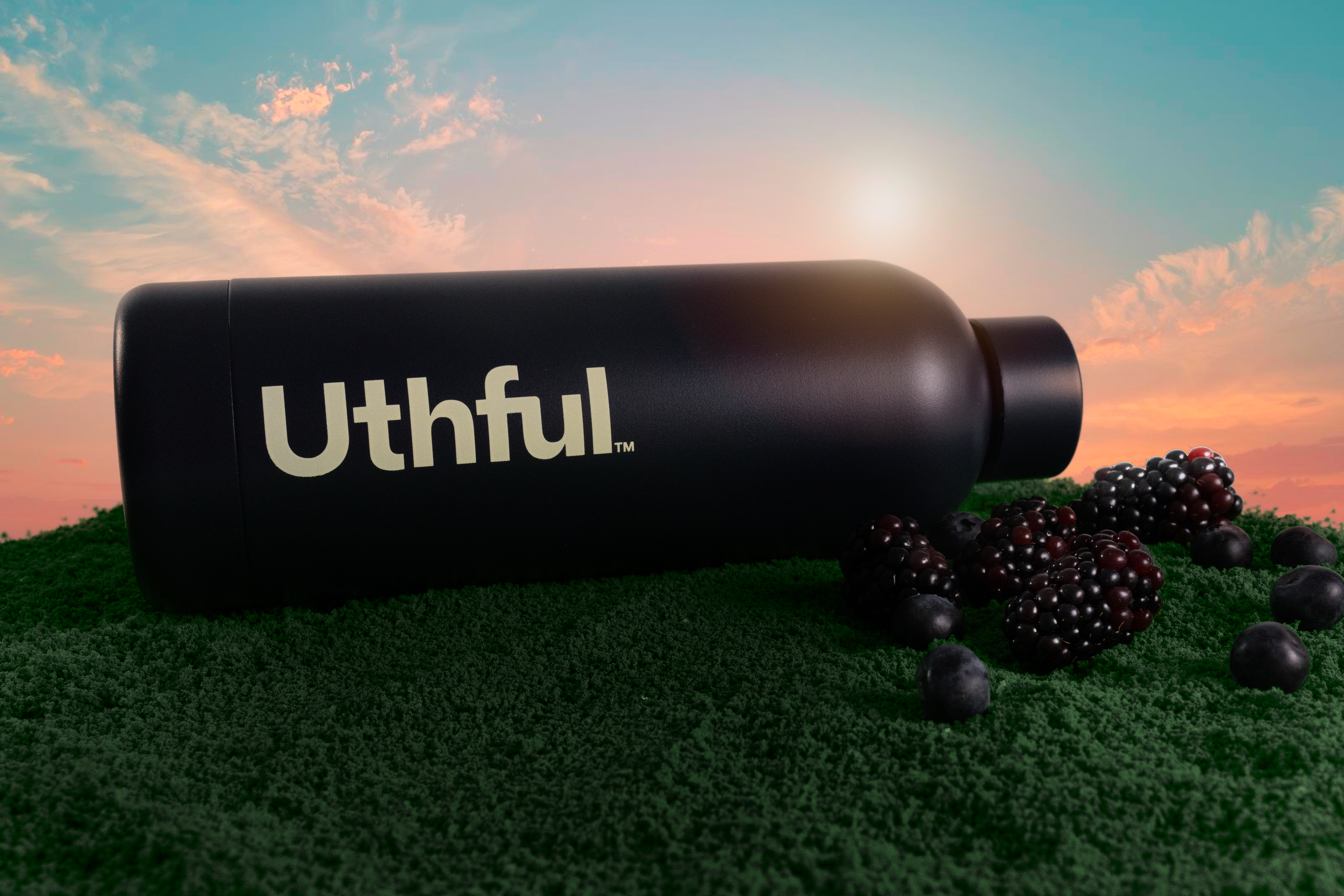 A black bottle labeled 'Uthful' lies on green grass with blackberries and blueberries scattered around, set against a sunset sky.
