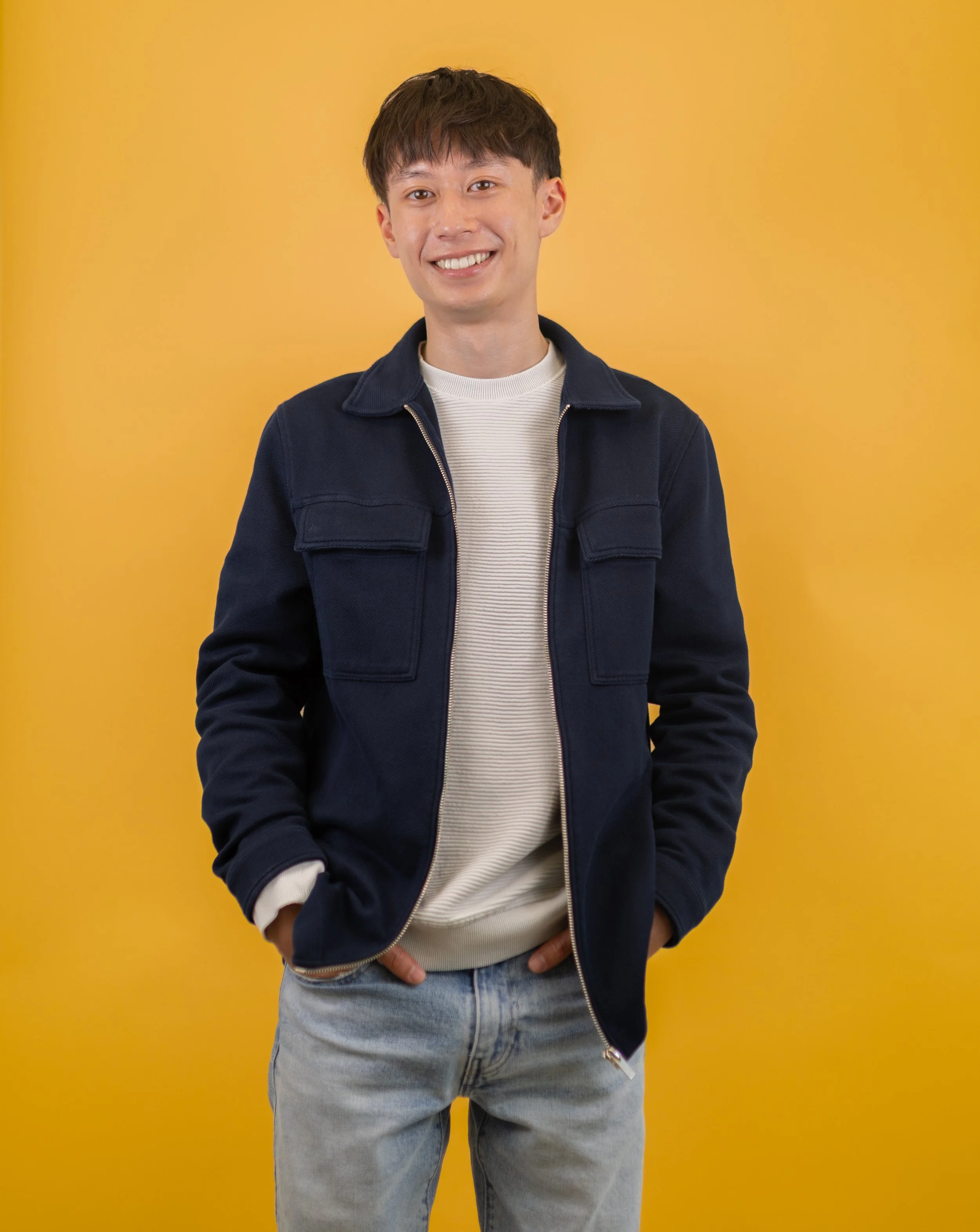 A young man with short dark hair and a friendly smile, wearing a navy blue jacket over a cream sweater and light blue jeans, standing with hands in pockets against a yellow background.