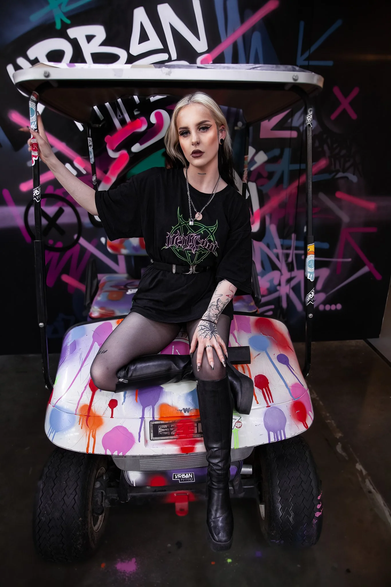 A young woman with blonde and black hair, tattoos, and dark makeup sitting on a graffiti-covered golf cart in front of a vibrant, graffiti-style background.