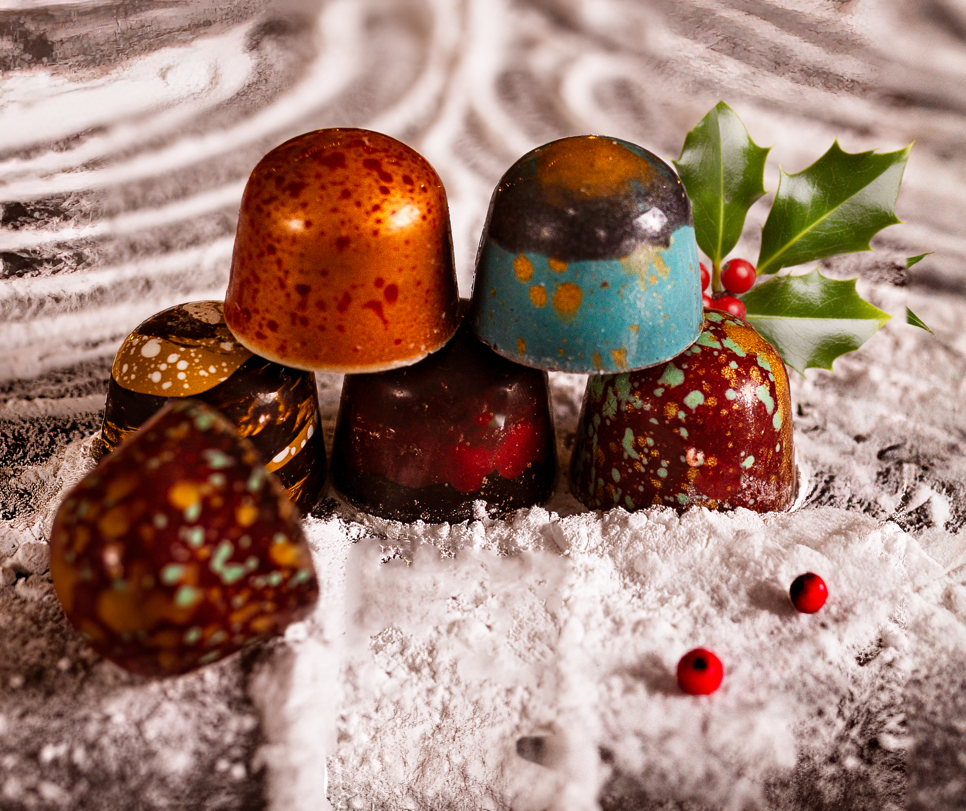 Decorative holiday chocolates arranged on white snow-like surface with holly leaves and red berries.