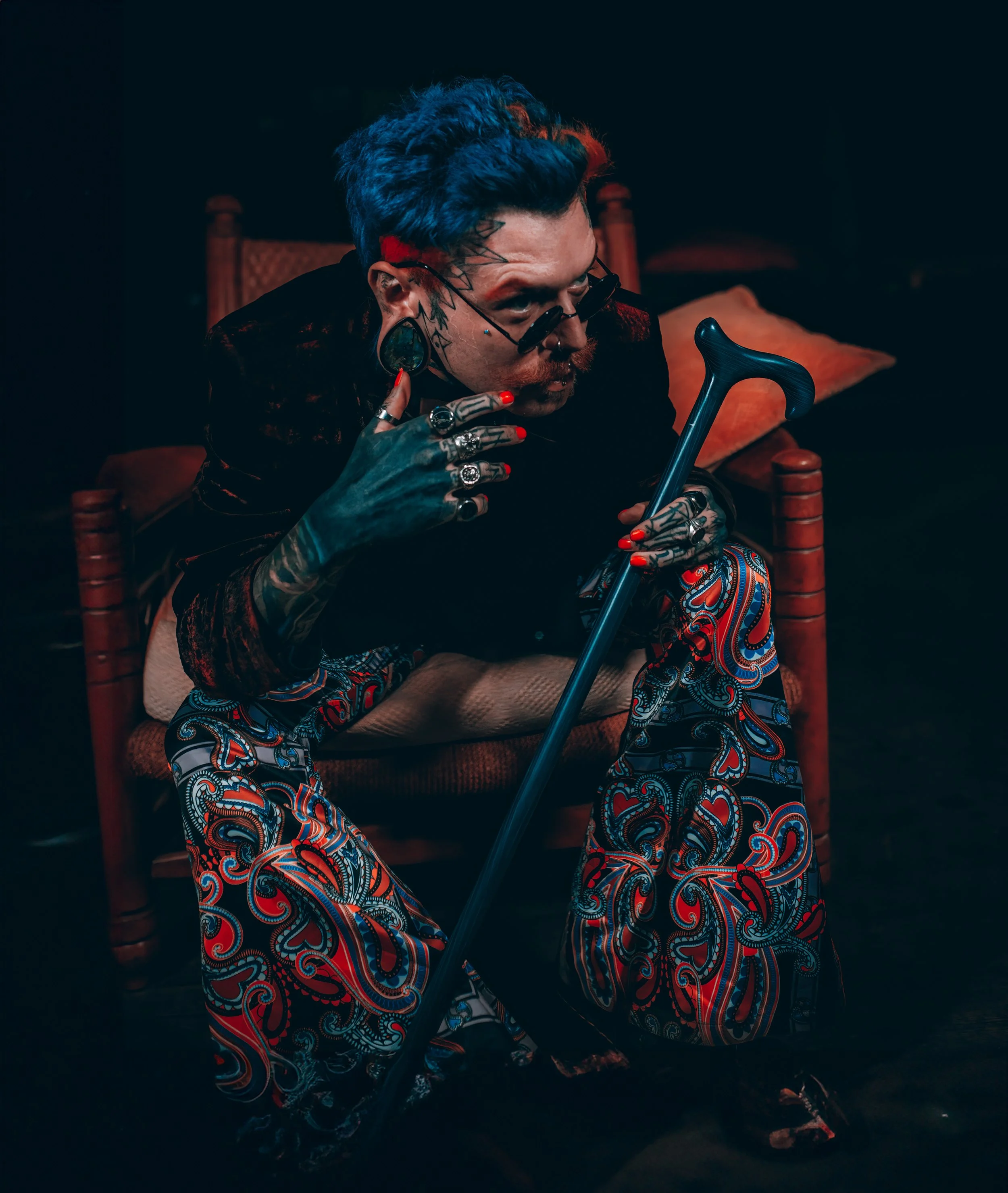 A man with colorful hair, tattoos, and accessories sitting on a wooden chair, holding a cane, wearing patterned pants and dark glasses, in a dimly lit room.