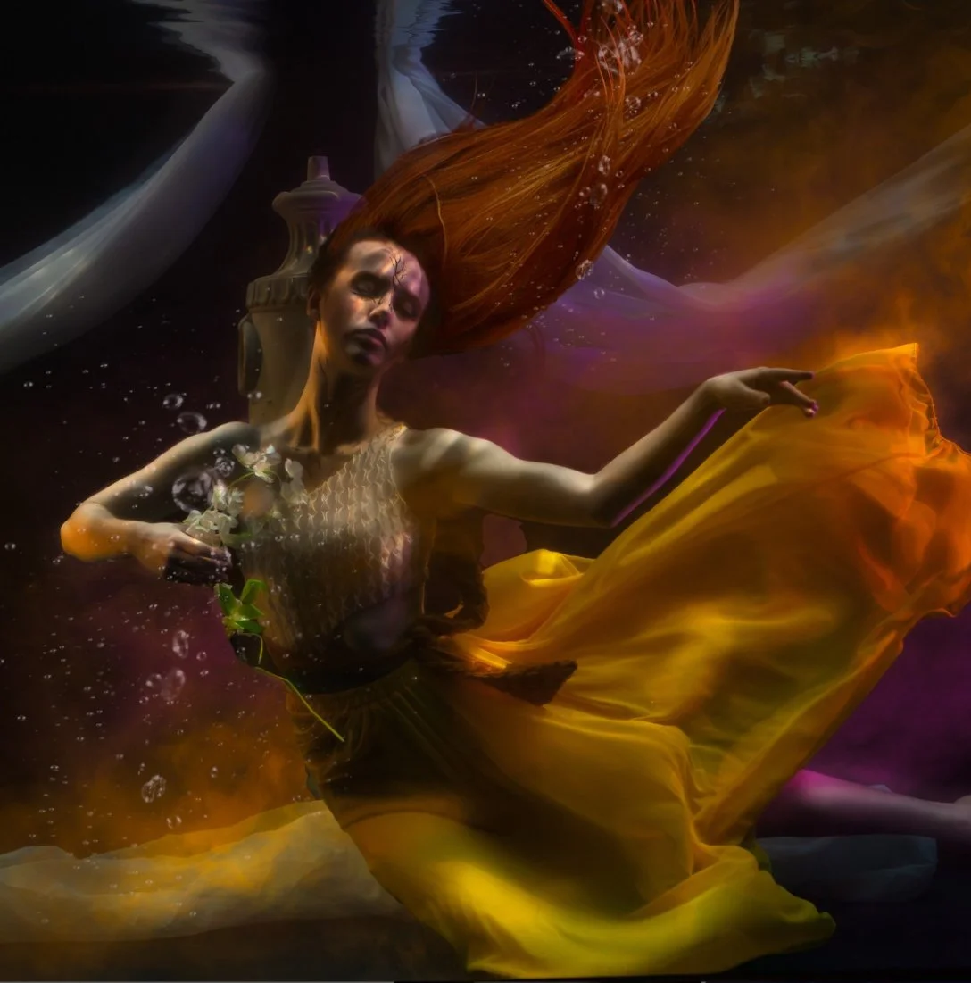 A woman with long red hair submerged underwater, wearing a golden dress and holding a branch with leaves, with water bubbles around her.