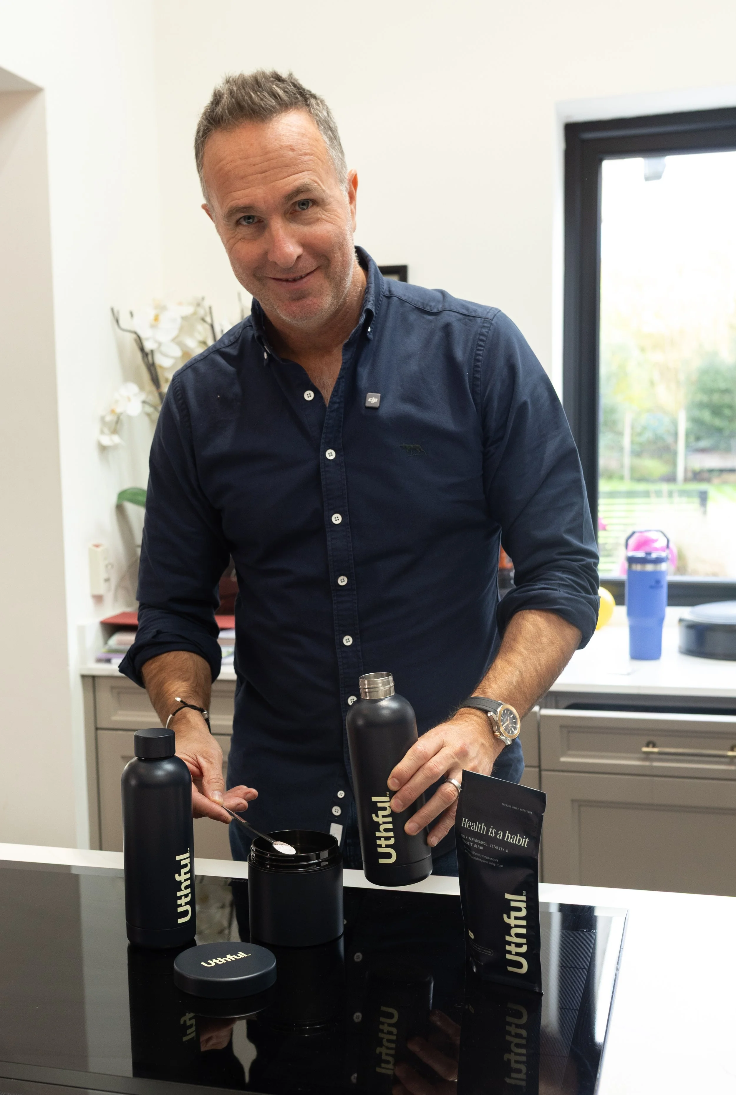 A man stands in a kitchen with a smile, holding a black water bottle. There are other black Utthful branded items, including a container, a packet, and a lid, on the black countertop. The man is wearing a dark blue shirt and a wristwatch, with a wind