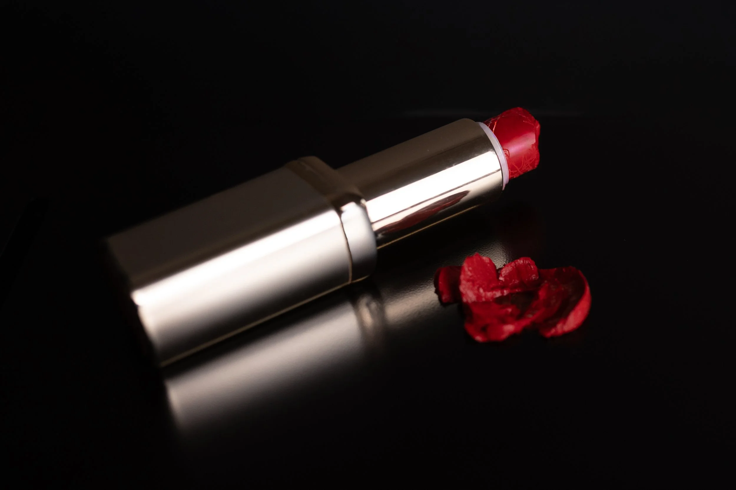 Commercial photography of a silver lipstick tube with red lipstick and red flower petals on a black reflective surface.