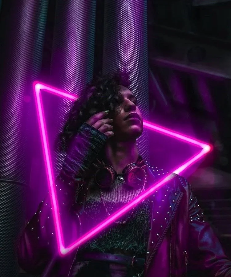 A woman with curly hair wears headphones around her neck and a leather jacket with studded details, standing against a metallic textured background illuminated by pink neon lights forming an angular shape.