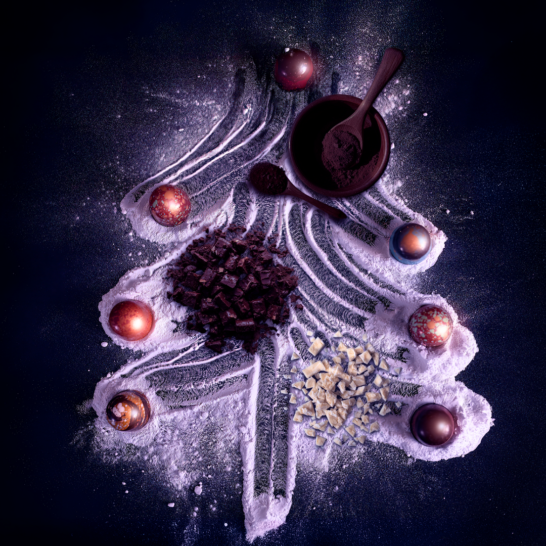 Festive holiday scene with Christmas ornaments, chocolate chunks, white chocolate pieces, and powdered sugar on a dark surface, arranged around a cocoa powder-filled bowl with a spoon.