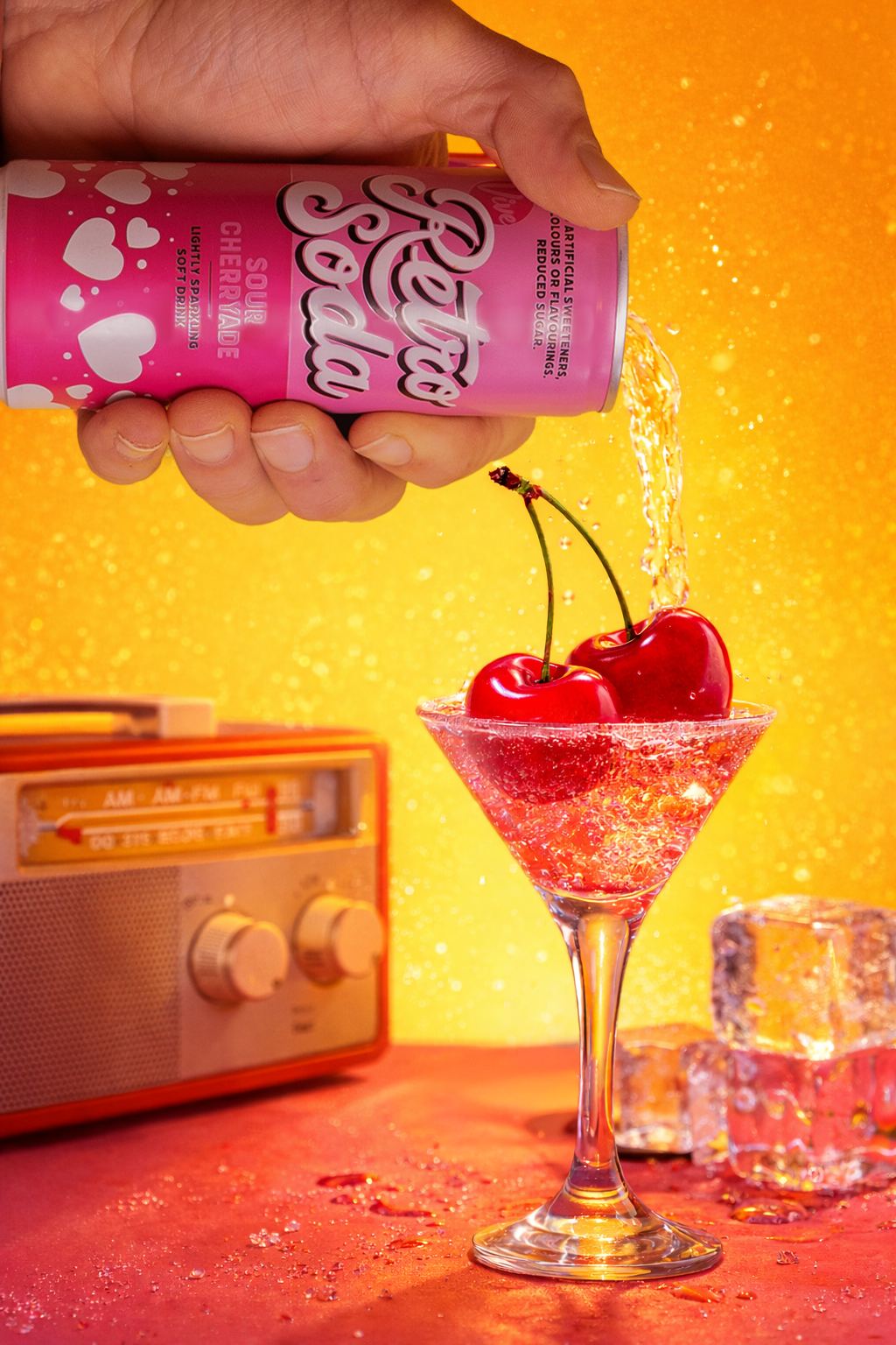 Still life photography showing a  hand pouring Fayrouz cherry soda from a pink can into a martini glass filled with ice and cherries, with a retro orange radio and a yellow backdrop.