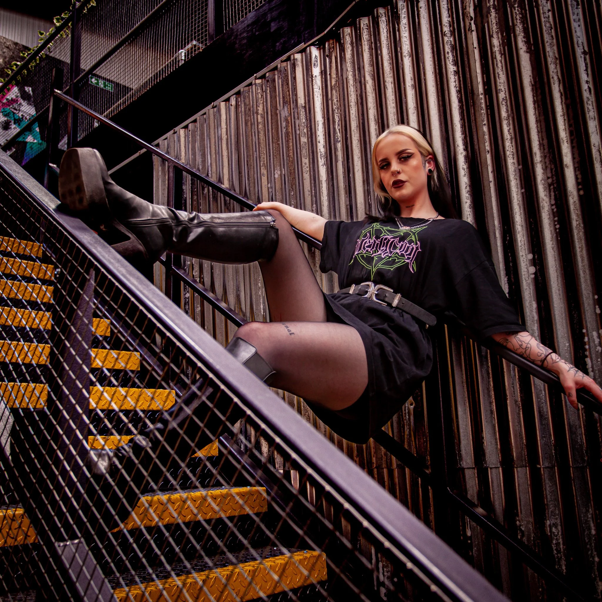 A young woman with blonde and black hair sitting on an industrial staircase with yellow steps, wearing a black band T-shirt, black shorts, sheer black tights, and black knee-high boots. She has tattoos and dark makeup, with an urban background of met