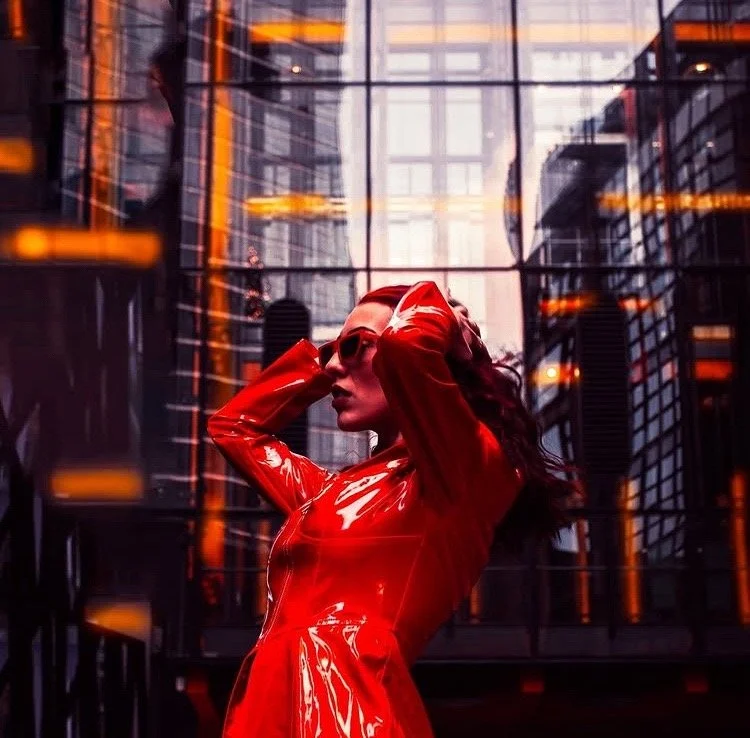 A woman in a shiny red dress and sunglasses posing in front of a glass building with reflections of city skyscrapers.