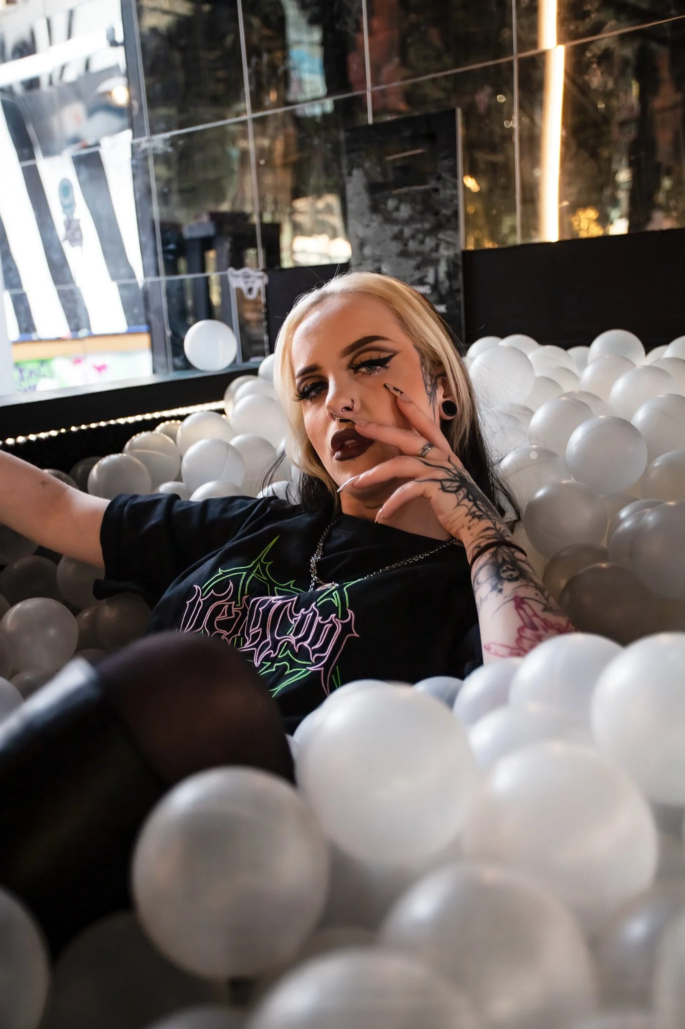 Young woman with platinum blonde and black hair, tattoos, dark makeup, wearing a black T-shirt with colorful graphics, lounging in a ball pit filled with white plastic balls.