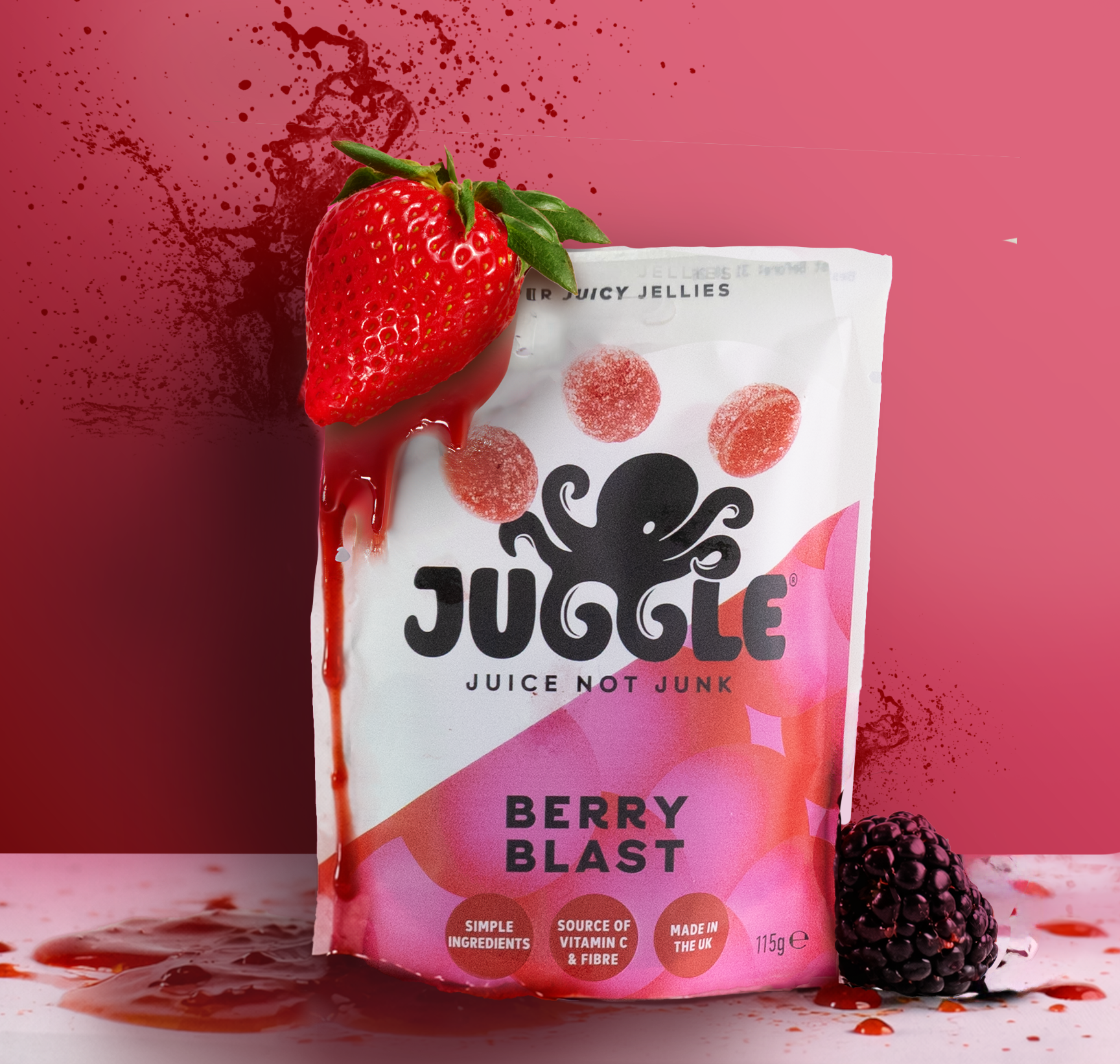 A pink background with a juice pouch labeled 'Juggle Berry Blast' musicemphasized with a black octopus logo, a strawberry with dripping juice perched on top, and a blackberry on the table with spilled juice and splatters.
