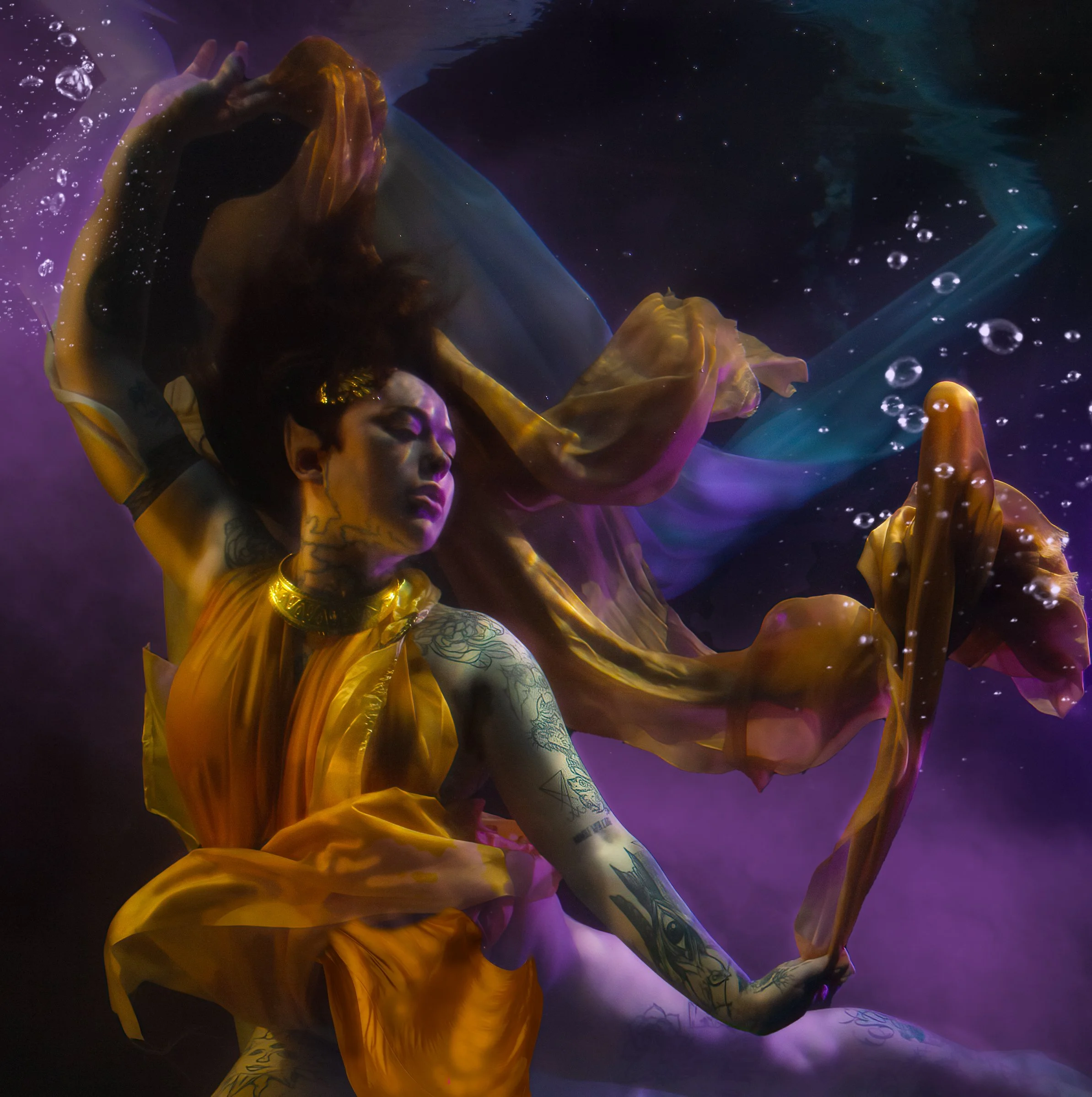 A woman with tattoos, wearing a yellow dress, is underwater, surrounded by purple and blue hues with bubbles. She appears to be posing or floating gracefully.