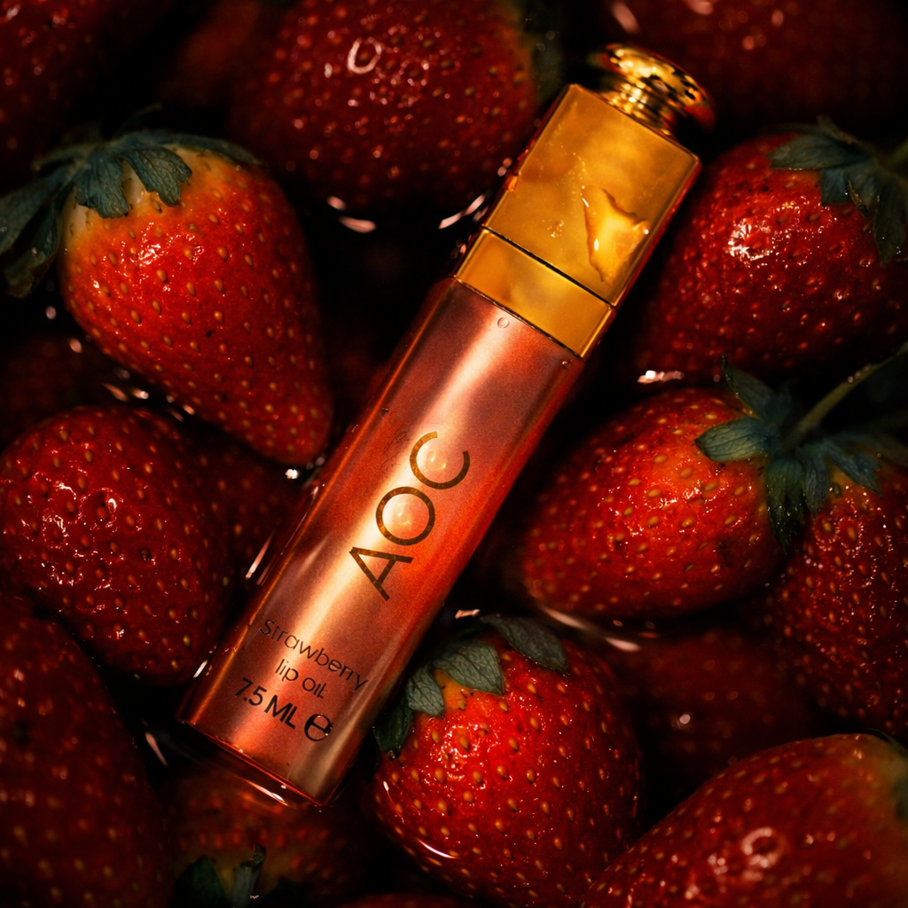 A rose gold tube of AOC strawberry lip oil surrounded by fresh strawberries.