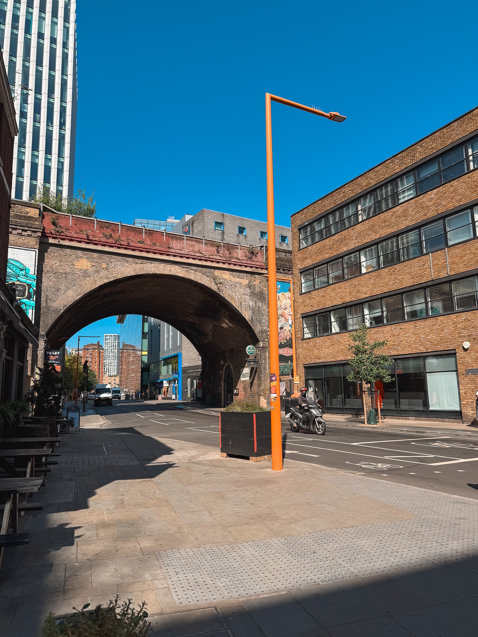 Empty city street with brick and glass buildings, underpass bridge, orange streetlamp, motorbike rider, and parked cars on a sunny day.