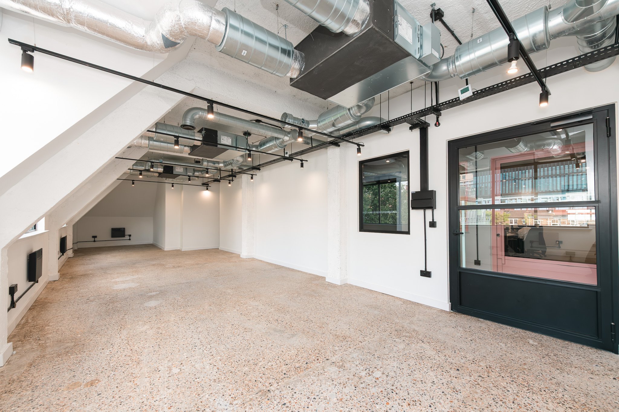 Empty modern room with white walls, exposed ventilation ducts, black track lighting, and large windows with black frames.