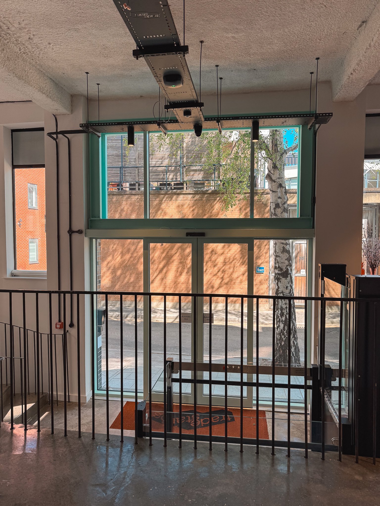 Indoor view of the entrance door to a building, with a metal railing, a security sensor at the top of the door frame, and a red doormat outside. The door is glass with a large window above, through which an outdoor scene with a tree, brick wall, and