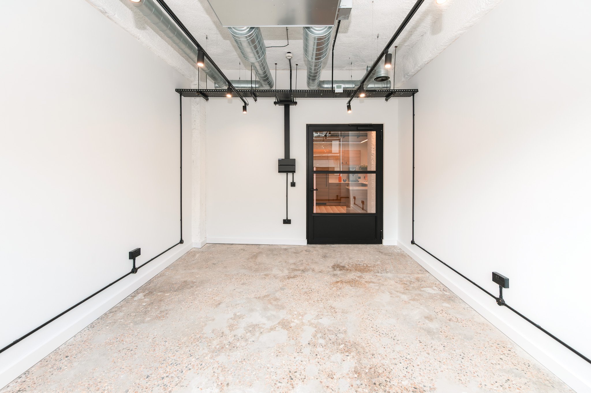 Empty room with white walls, exposed ceiling ducts, black electrical conduit and outlet boxes, and a black door with glass panel leading to another space.