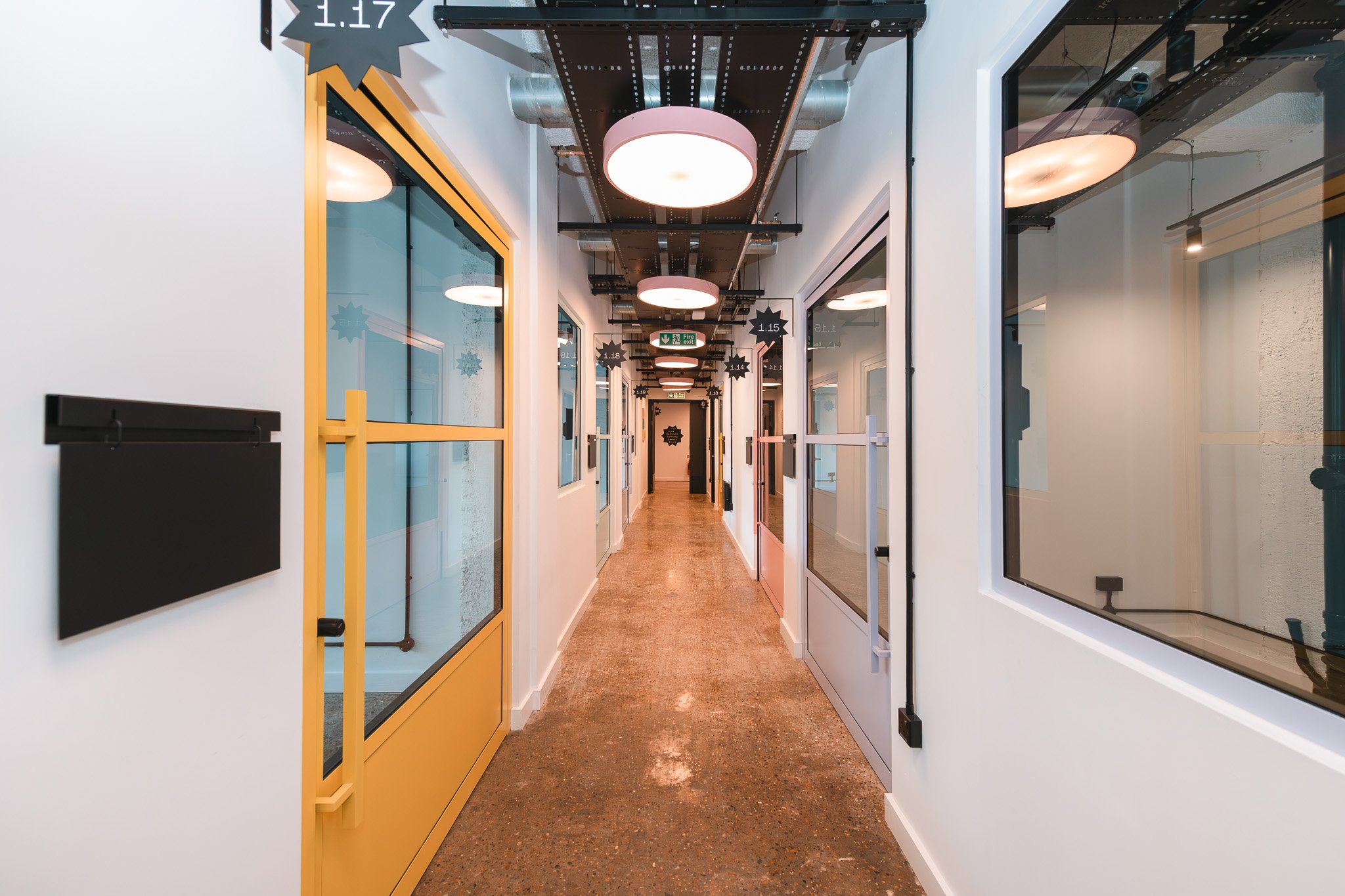 Interior office hallway with white walls, colorful doors, ceiling lights, and signs indicating room numbers.