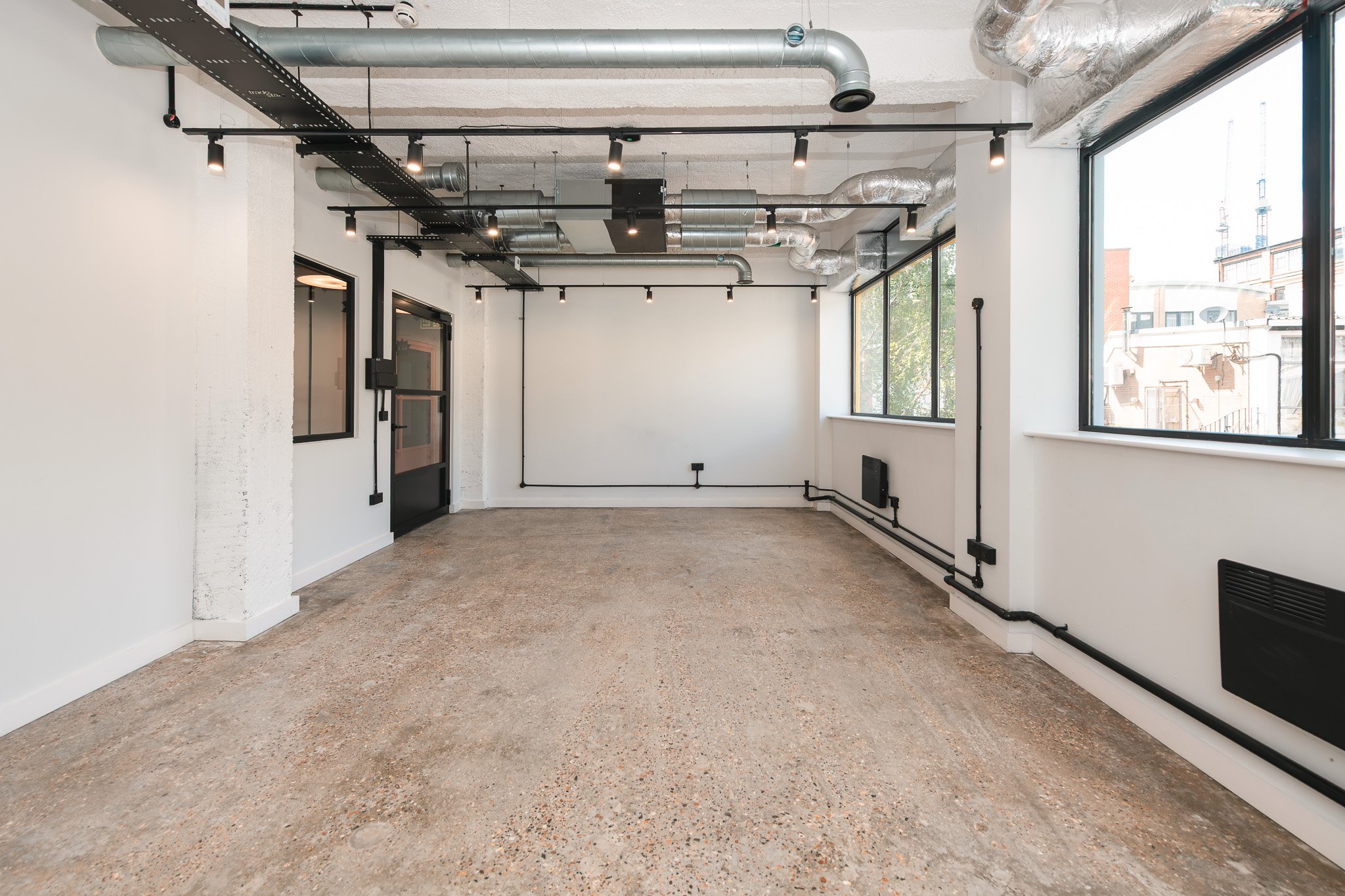 Empty room with large windows, exposed ductwork on ceiling, and black piping along walls.