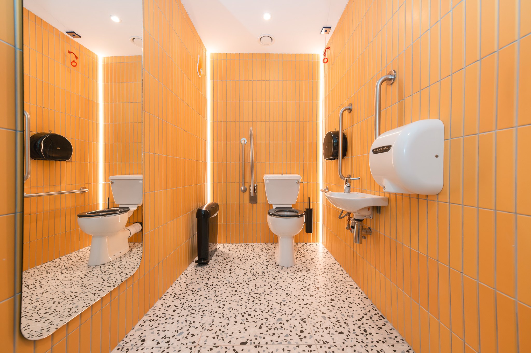 Accessible public restroom with orange tiled walls, a toilet, a sink, grab bars, a mirror, and a paper towel dispenser.