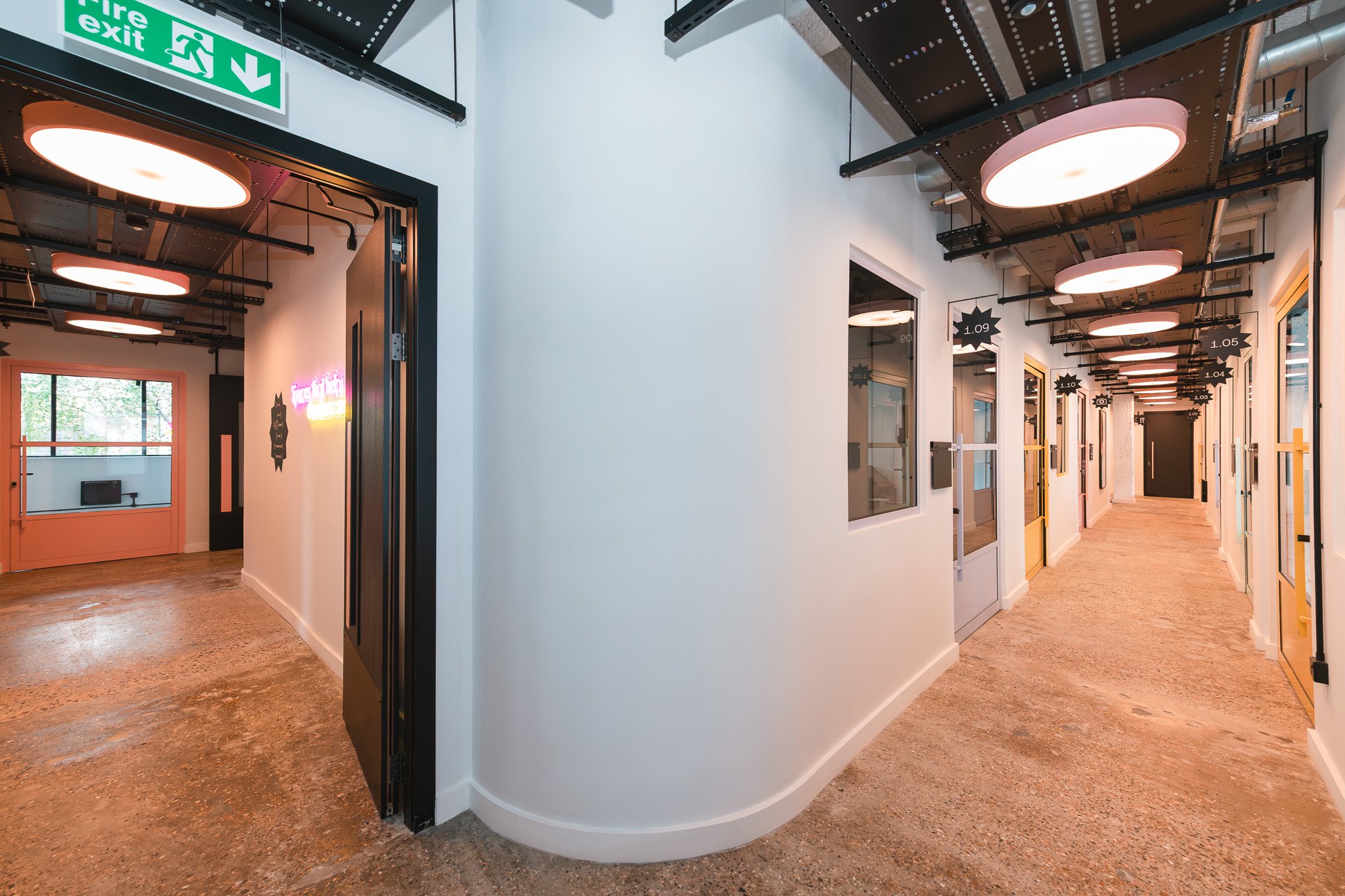 Interior hallway with conference rooms on the right side, some with yellow doors, and a fire exit sign at the ceiling. Modern ceiling lights illuminate the space and windows provide natural light.