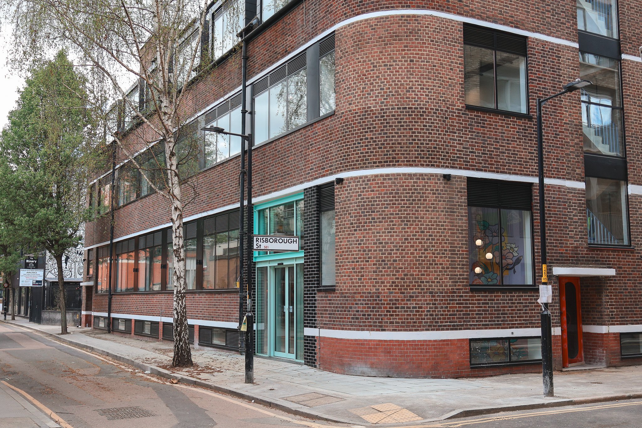 A street corner with a modern brick apartment building on Risborough Street, with large windows, trees, street lamps, and a sidewalk.