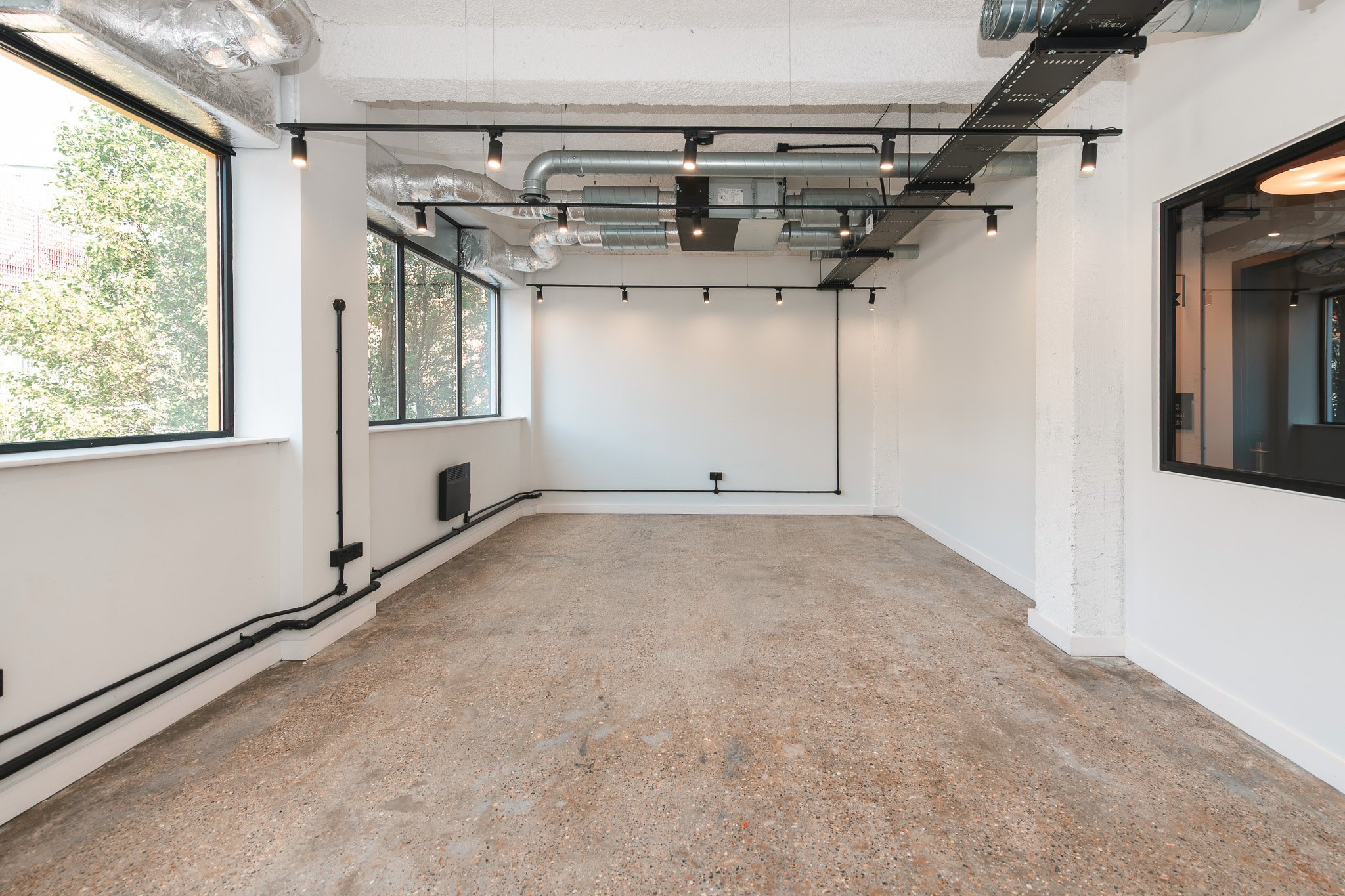 Empty modern room with large windows, exposed ductwork, black track lighting, and a concrete floor.